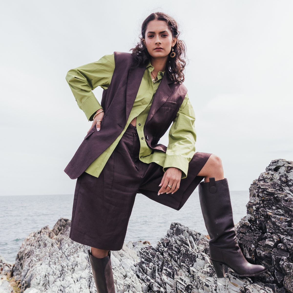 A model stands on rocky coastal ground wearing a sage green shirt, a dark brown structured vest, a matching dark brown skirt, and dark brown knee-high boots.