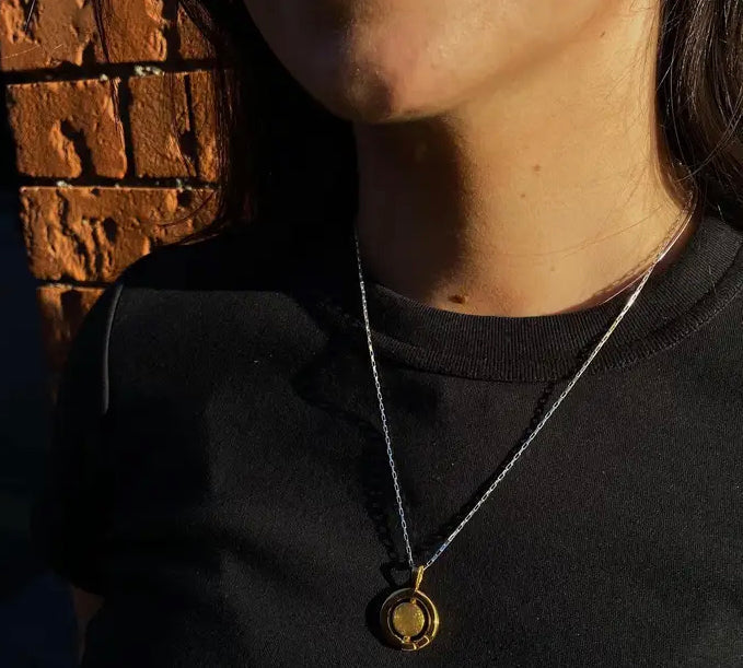 A person wearing a black shirt stands against a textured brick wall. They have a Gallardo & Blaine Designs Urú Large Pendant & Chain hanging around their neck. The sunlight casts shadows, partially illuminating their face and chest.