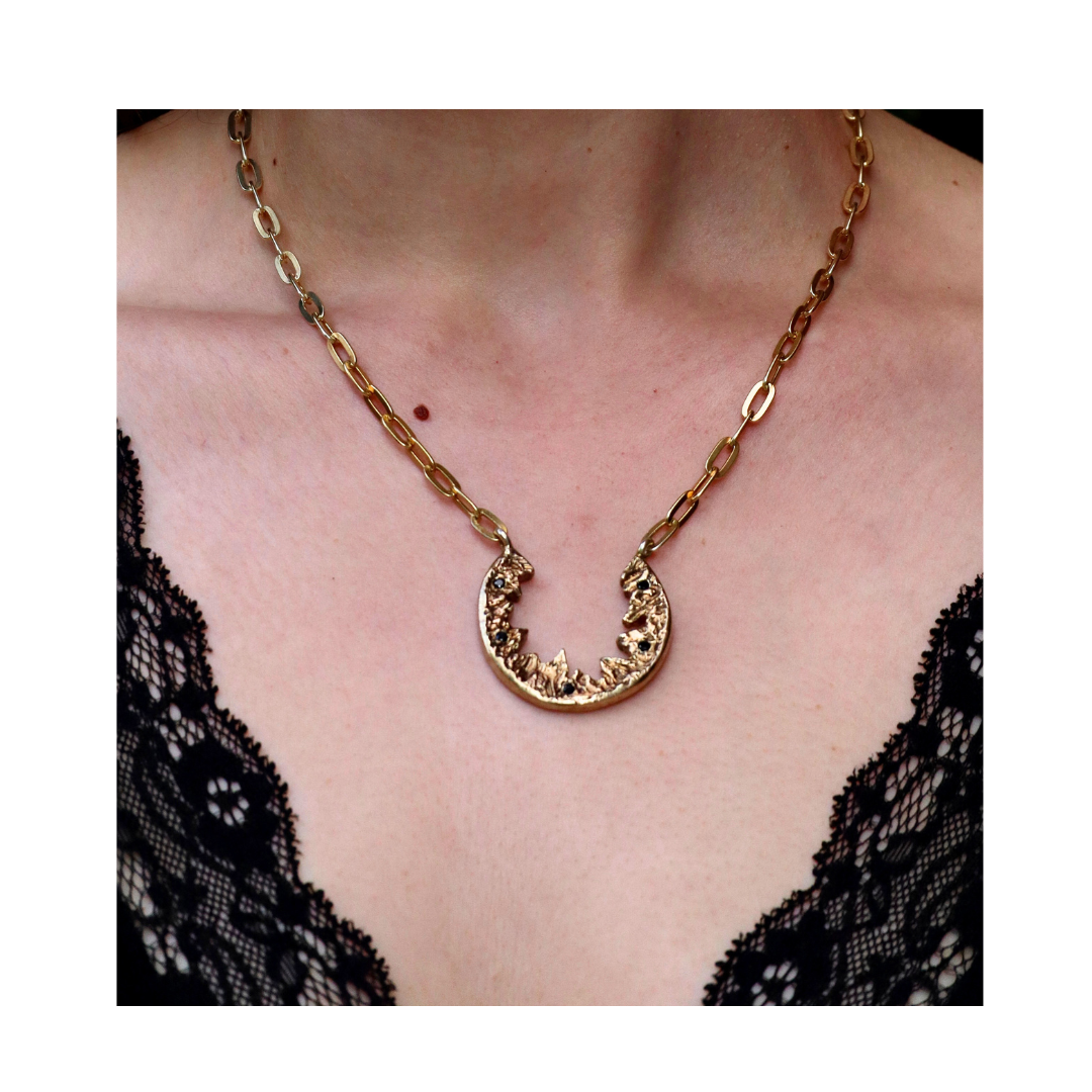 A person wears the Madeleine Blaine Elements Necklace—featuring a gold chain and a crescent-shaped, hand-carved textured pendant—resting against bare skin and a black lace-trimmed garment; an exquisite piece of nature-inspired jewelry.