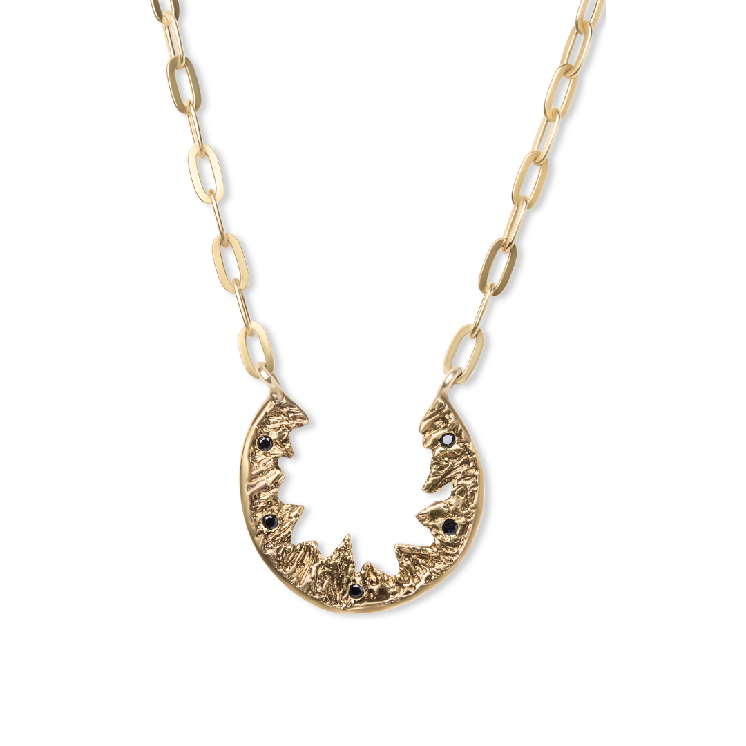 The Elements Necklace by Madeleine Blaine features a gold chain with elongated links and an oval, jagged pendant adorned with hand-carved textured details and small dark stones, capturing the essence of nature-inspired jewelry in the brand’s signature sty