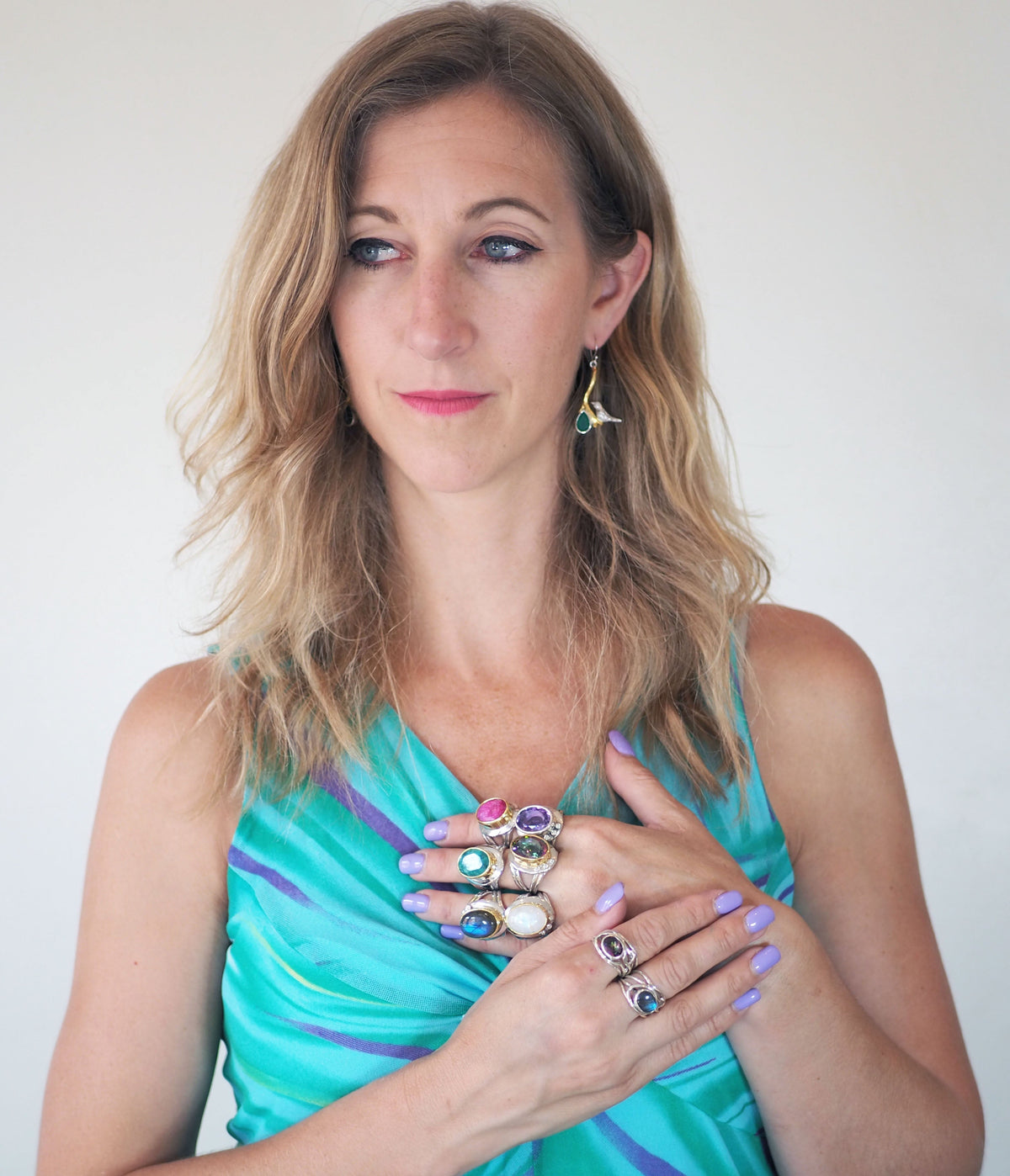 A woman with blonde hair is wearing a turquoise and blue dress. She has several colourful Eyetelia Rings in various gemstones by Gallardo & Blaine Designs on her fingers, their vibrant hues matched perfectly by her nail polish. Adorning her ear with a pen