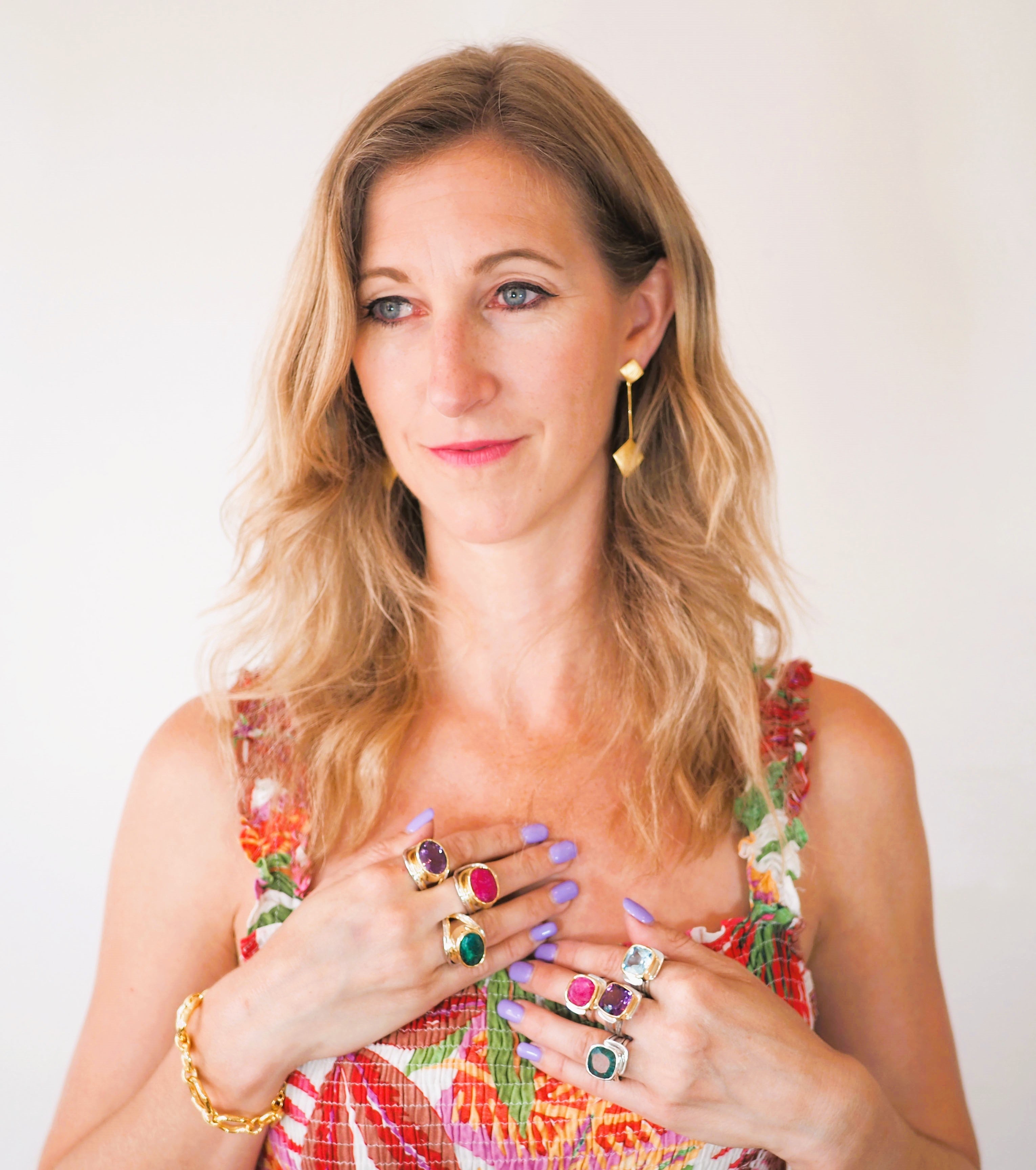 A person with wavy blonde hair is wearing a colorful floral top. They are adorned with multiple rings with large, colorful stones on both hands, along with a gold vermeil bracelet and Art Deco Earrings In Gold Vermeil by Gallardo & Blaine Designs featurin