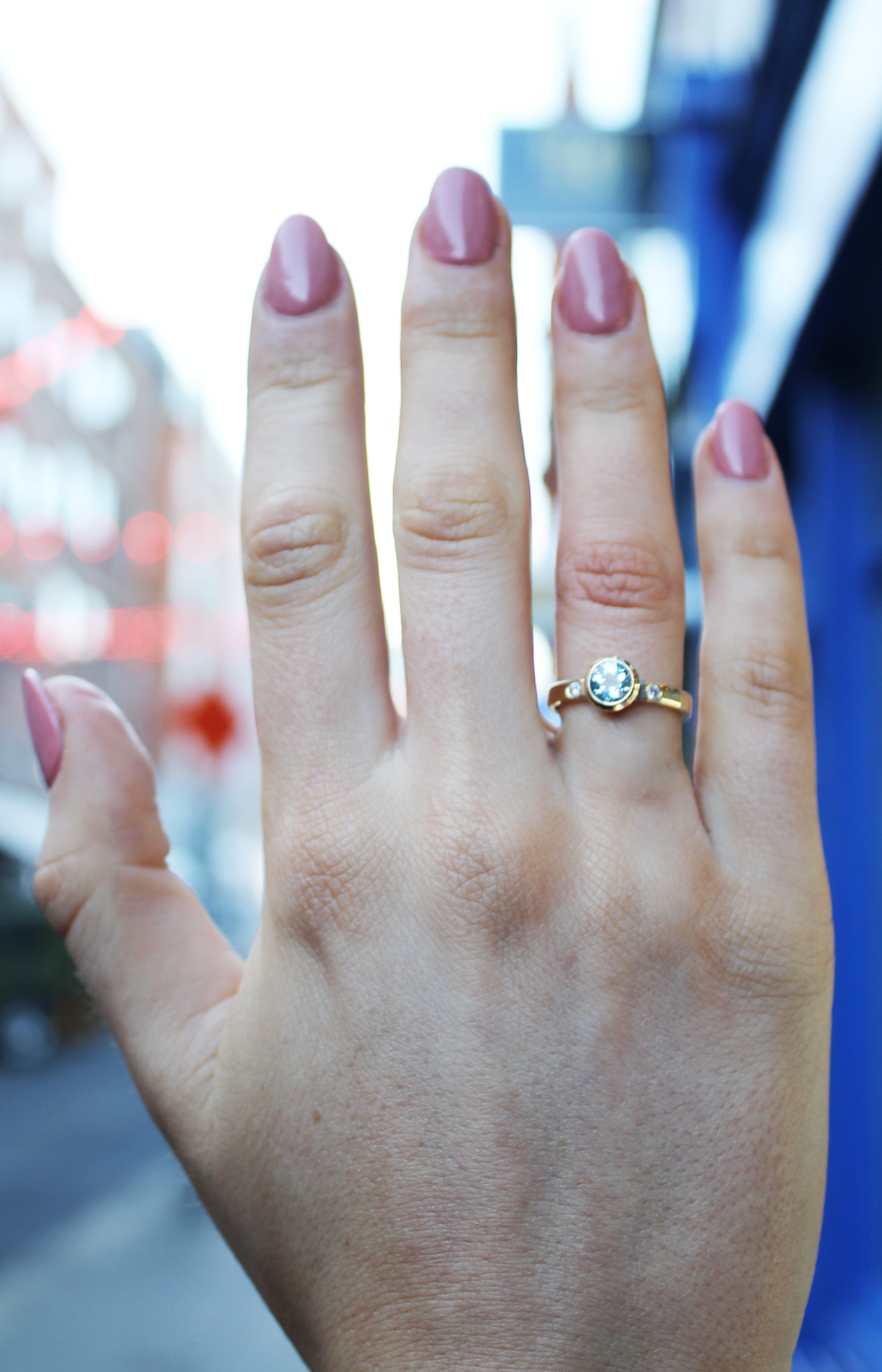 A hand with pink-painted nails displays the Timeless Cascade 9ct Yellow Ring by Madeleine Fine Jewellery, featuring a round clear gemstone on the ring finger, with an out-of-focus street scene in the background.
