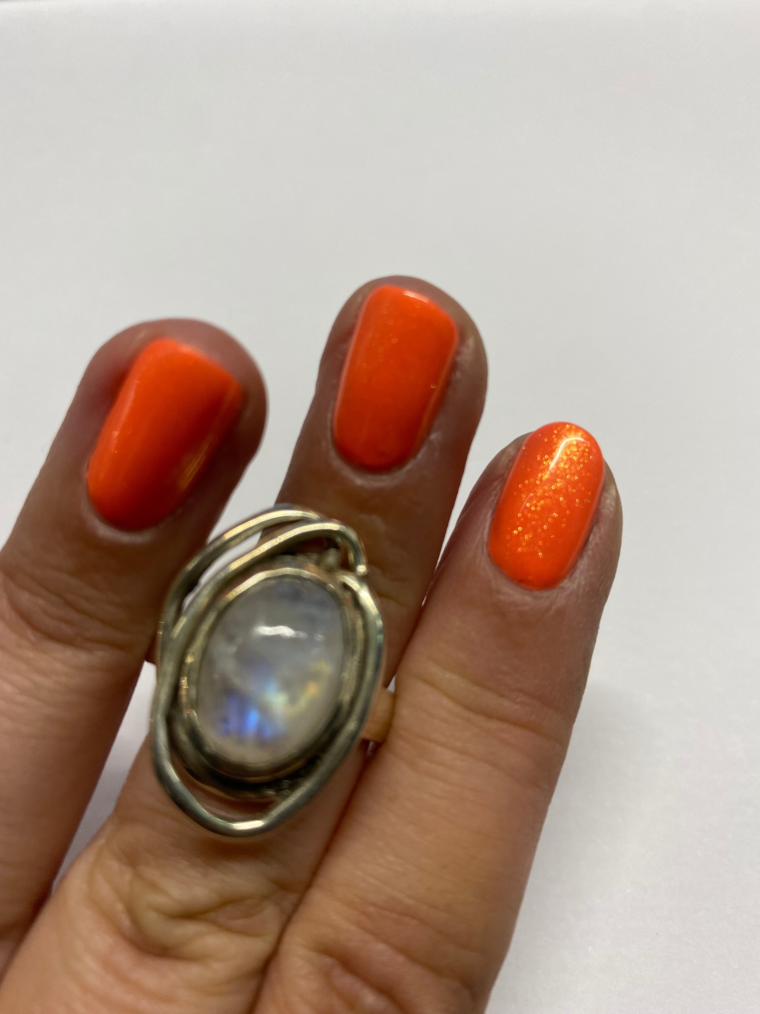 A hand with bright orange, shiny nail polish is shown. The ring finger wears a large, silver Gallardo & Blaine Designs Cosmos Ring Small In Various Stone featuring an oval-shaped milky white gemstone with a hint of blue iridescence. The nails are neatly m