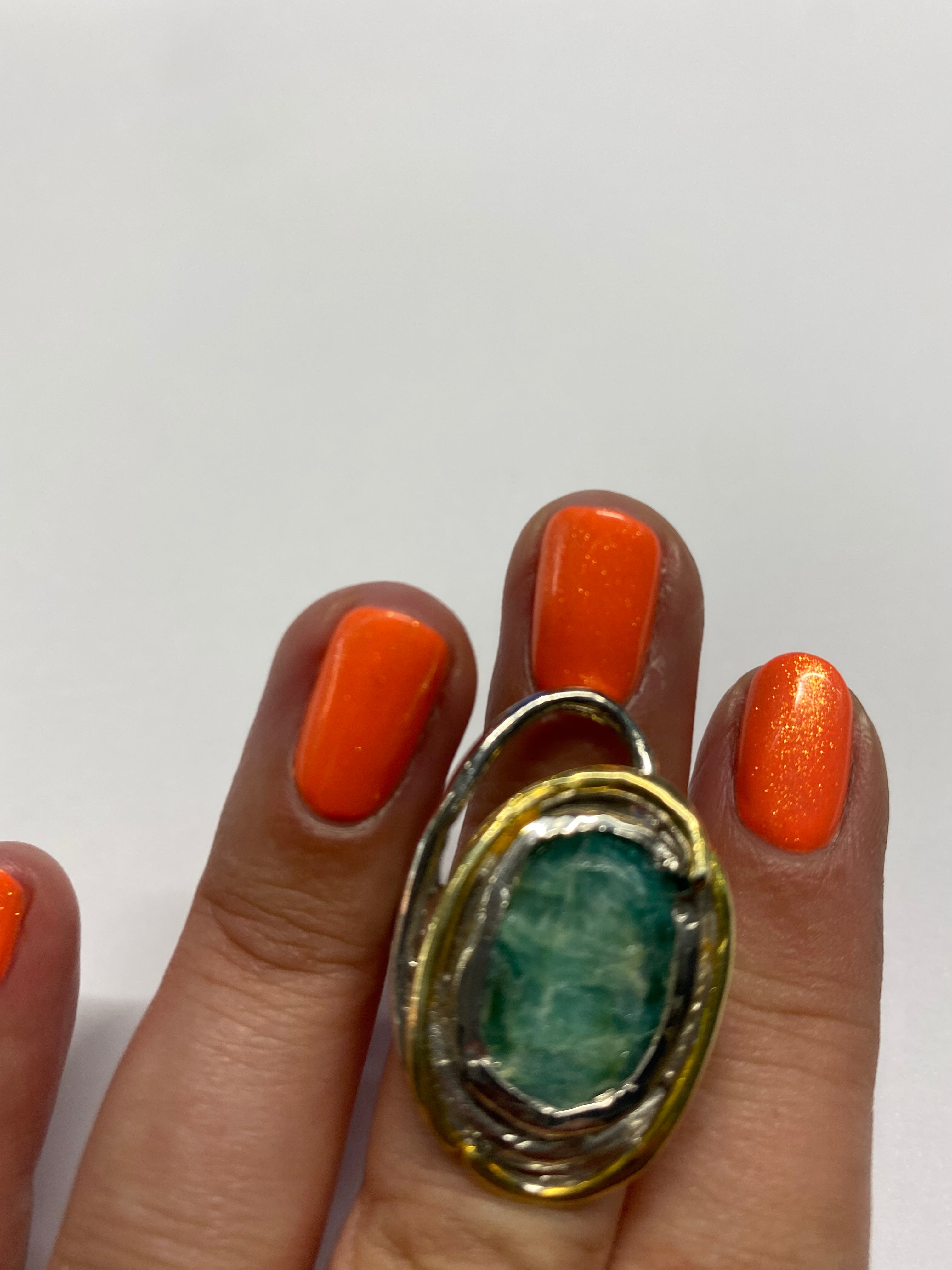 A hand with bright orange nail polish is shown wearing a Gallardo & Blaine Designs Cosmos Ring Large in Various Stones on the middle finger. The ring features a green gemstone set in an ornate, multi-layered band with gold plate accents. The background is