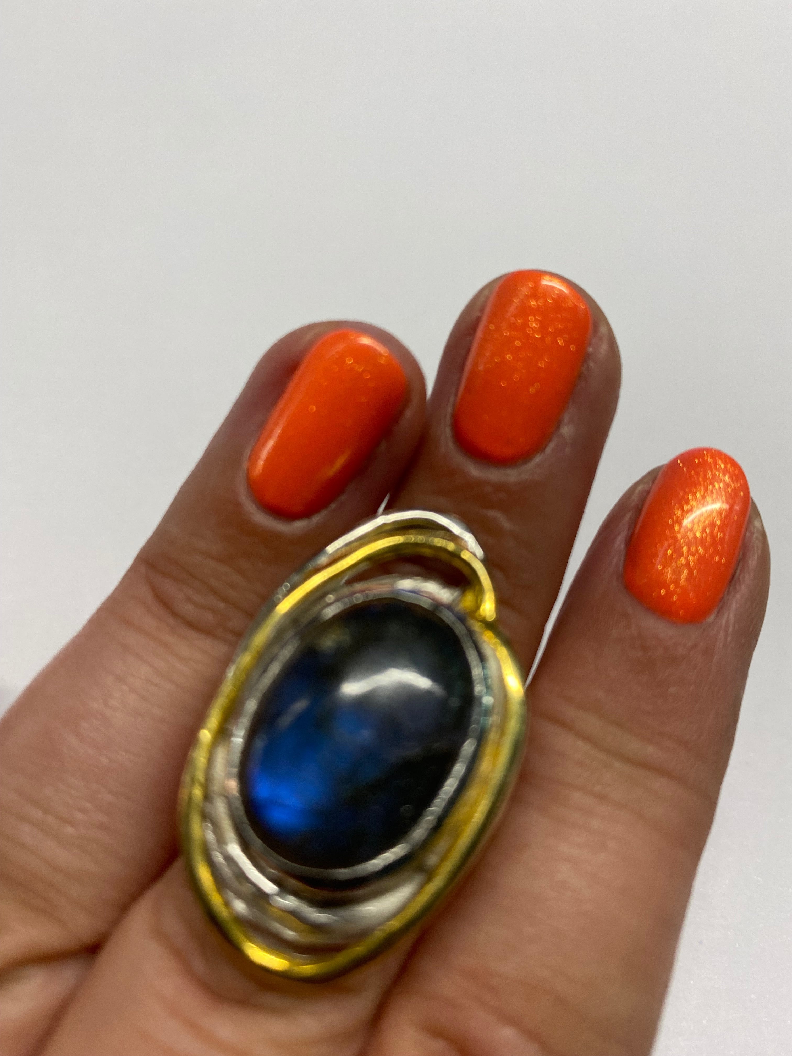 A close-up of a hand with bright orange painted nails, showcasing a stunning Cosmos Ring Large in Various Stones by Gallardo & Blaine Designs with a dark blue oval-shaped stone set in a silver and gold-toned setting. The background is plain white.