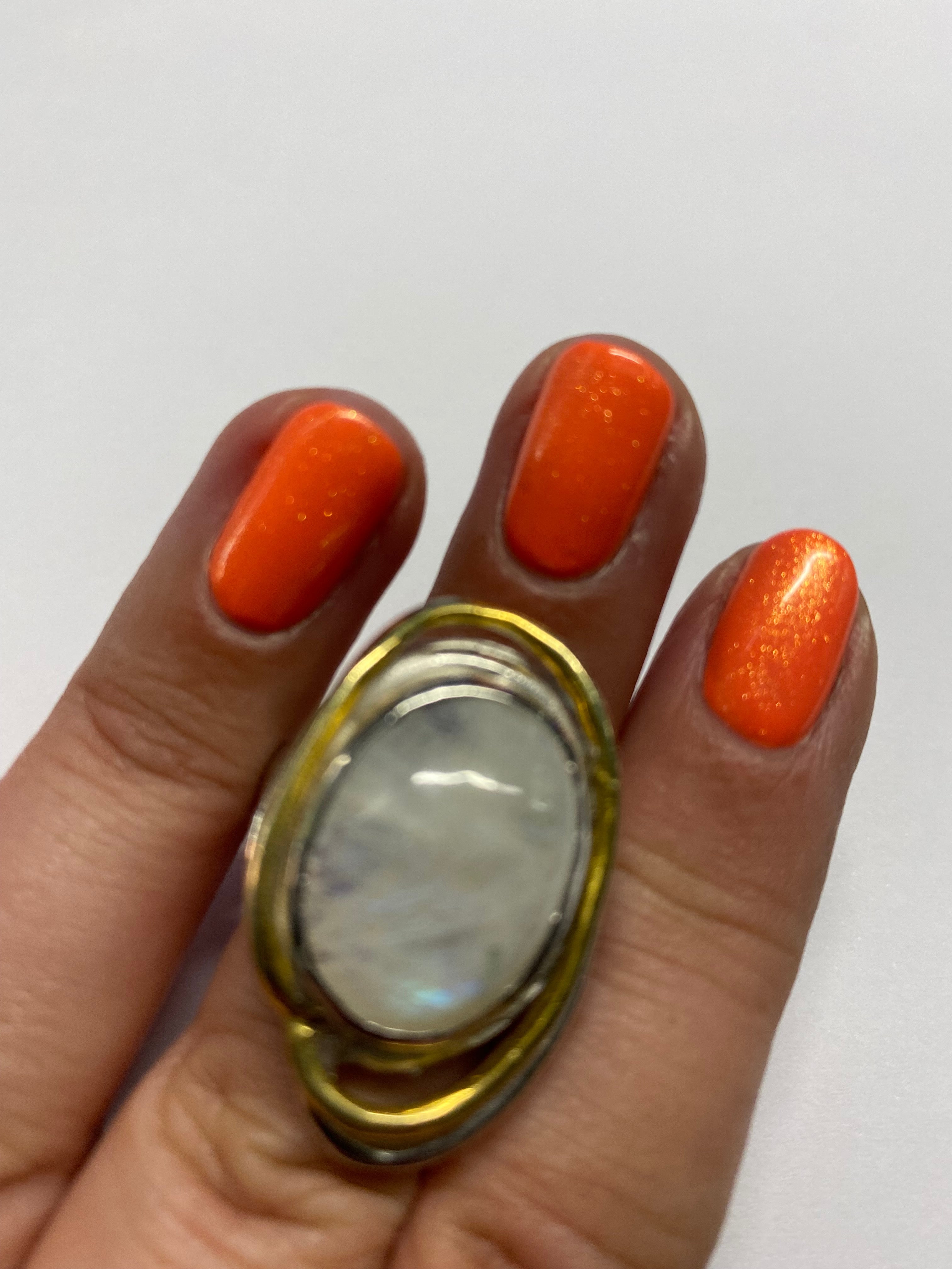 A hand with an orange glitter nail manicure is shown. The index finger is adorned with a Cosmos Ring Large in Various Stones by Gallardo & Blaine Designs, set in a gold and silver band. The background is plain white.