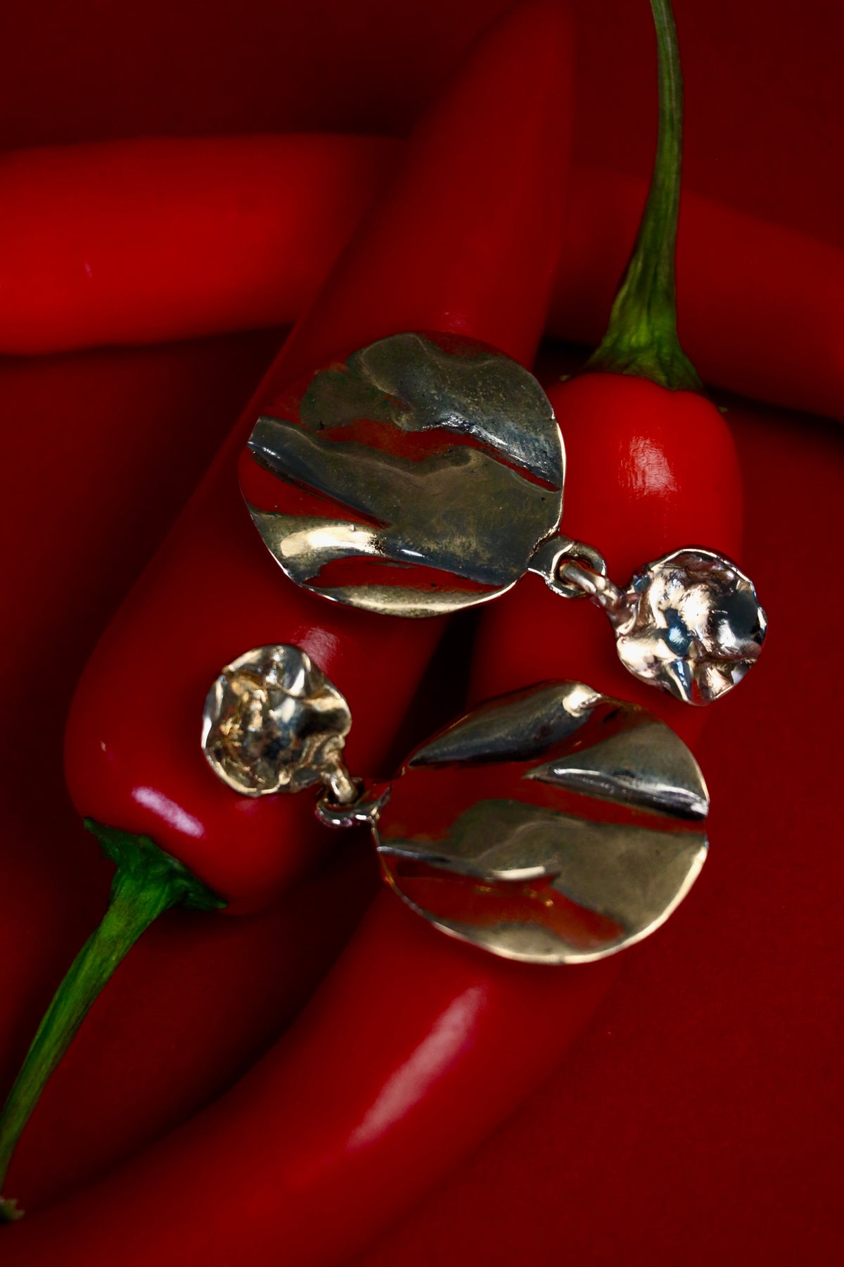 Two silver, abstract-shaped Tide Drop Earrings by Madeleine Blaine—hand-carved in Dublin—are displayed atop two vibrant red chili peppers against a matching red background, showcasing the contemporary charm of Irish jewelry.