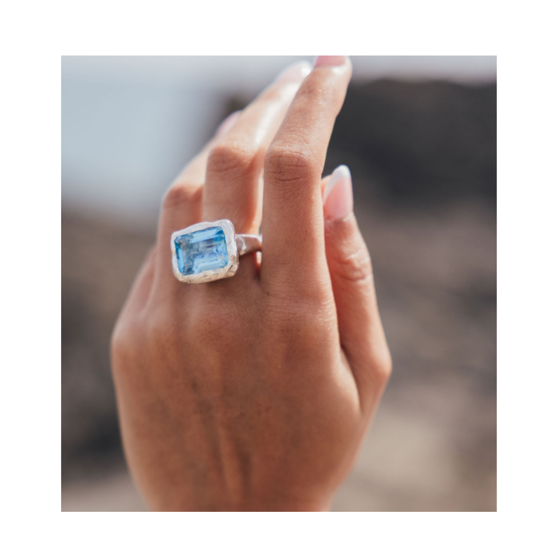 A hand adorned with the Glacier Ring by Madeleine Sculptural Jewellery, featuring a large rectangular blue gemstone set in silver, held up against a blurred outdoor background.