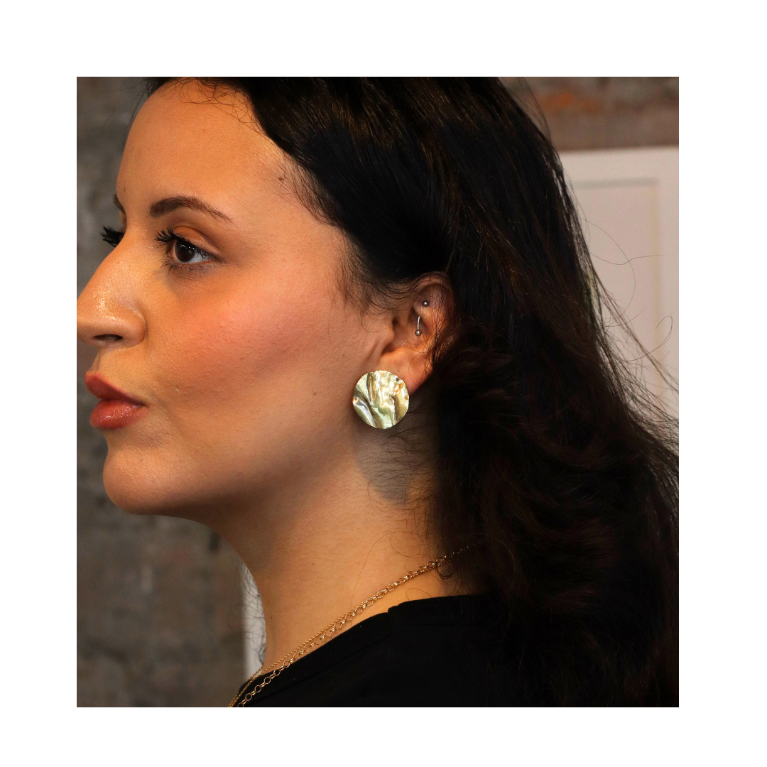 A woman with dark hair is shown in profile against a neutral background, wearing the Tide Studs Large earrings by Madeleine Blaine—hand-carved, round, iridescent jewelry inspired by the Irish coastline.