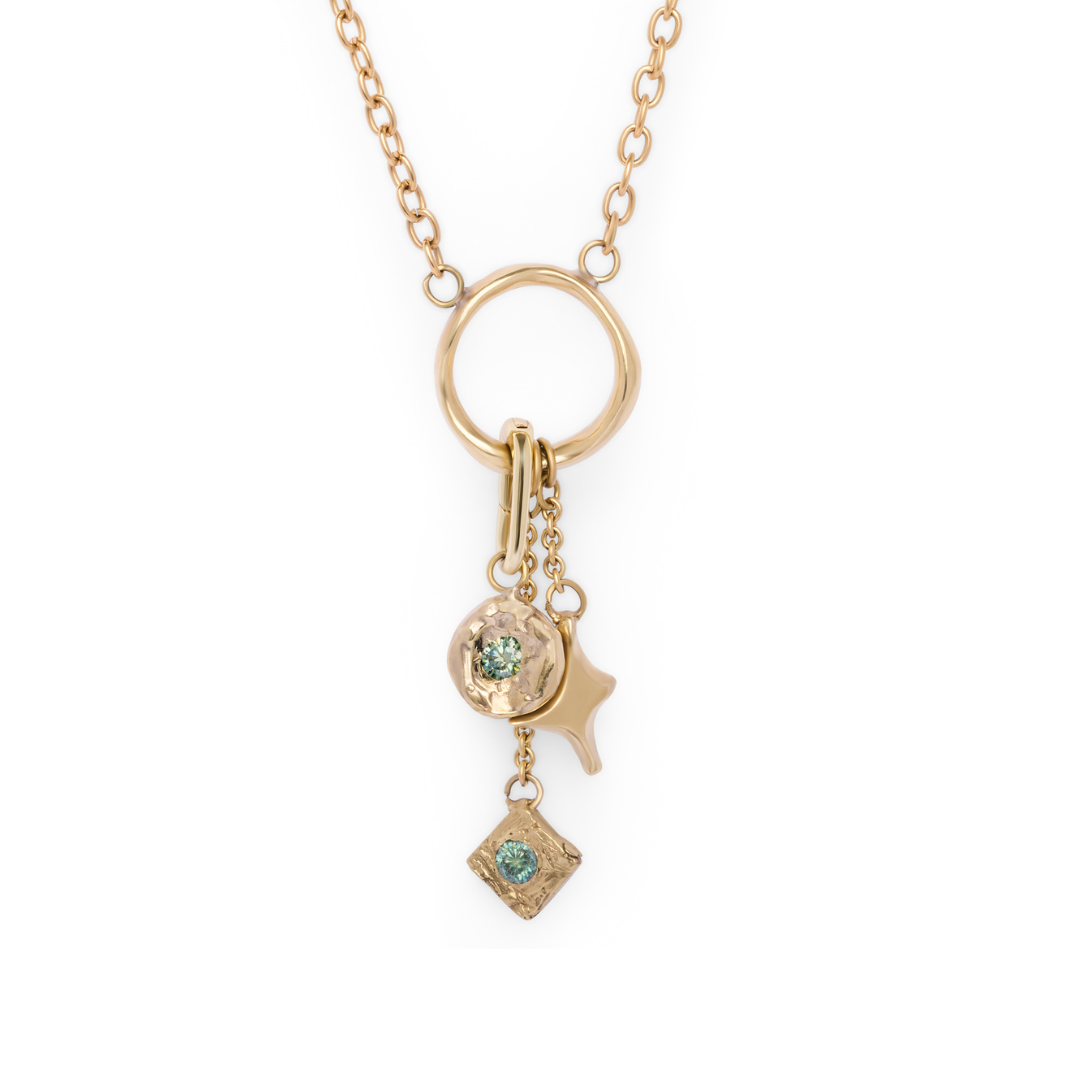 The Spark Necklace from Madeleine Sculptural Jewellery features a gold chain with a circular pendant, adorned with three dangling charms: a textured disc set with a green gem, an abstract star shape, and a small square charm accented by a green stone. Dis