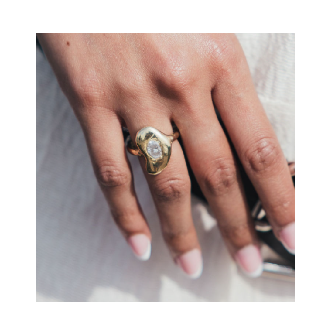 A hand adorned with the Carraig Ring from Madeleine Sculptural Jewellery—with its gold band and clear stone centerpiece—rests gracefully on a white fabric surface. The nails are manicured with a natural pink base and white tips, creating an elegant look i