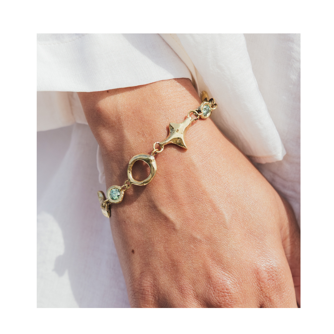 A close-up of a hand wearing the Spark Bracelet in Brass by Madeleine Sculptural Jewellery, featuring circular links and light blue gemstones, against a white fabric background.
