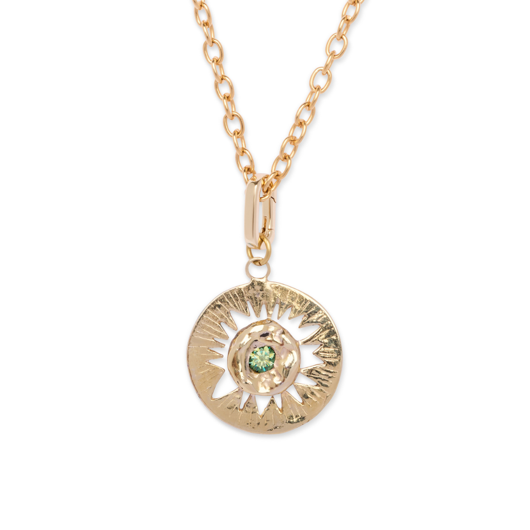The Tundra Necklace by Madeleine Sculptural Jewellery features a gold chain and a round, textured pendant adorned with a sunburst design and a green gemstone at its center.