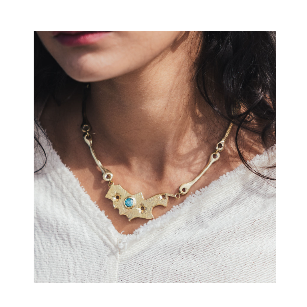 A woman wearing the Blaze Necklace by Madeleine Sculptural Jewellery—a textured, gold statement piece with an irregular shape and a blue gemstone—paired with a white V-neck top.