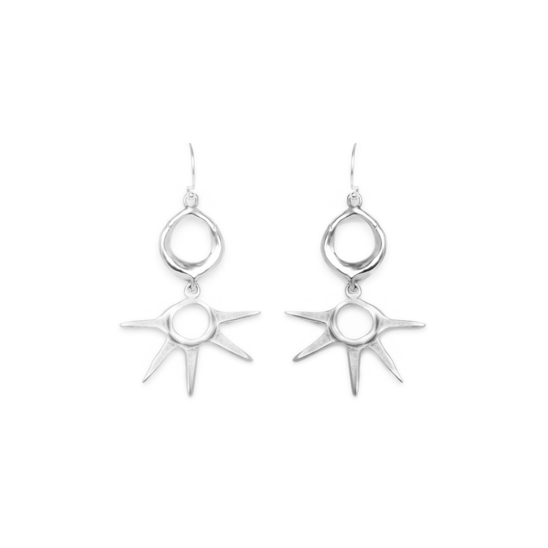 Introducing the Réalta Earrings by Madeleine Blaine—a pair of silver dangle earrings featuring oval loops at the top and striking sunburst designs with pointed rays suspended below, elegantly displayed on a white background. These luxury accessories showc