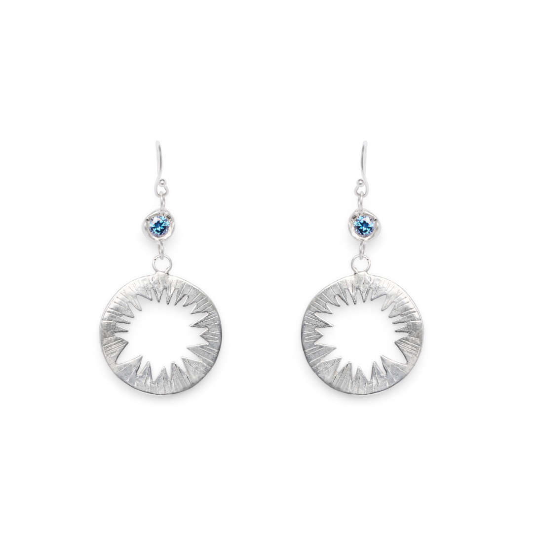 Tundra Earrings with Moissanite by Madeleine Blaine, featuring silver sunburst circle pendants and a small blue moissanite gemstone above each pendant, displayed against a white background.