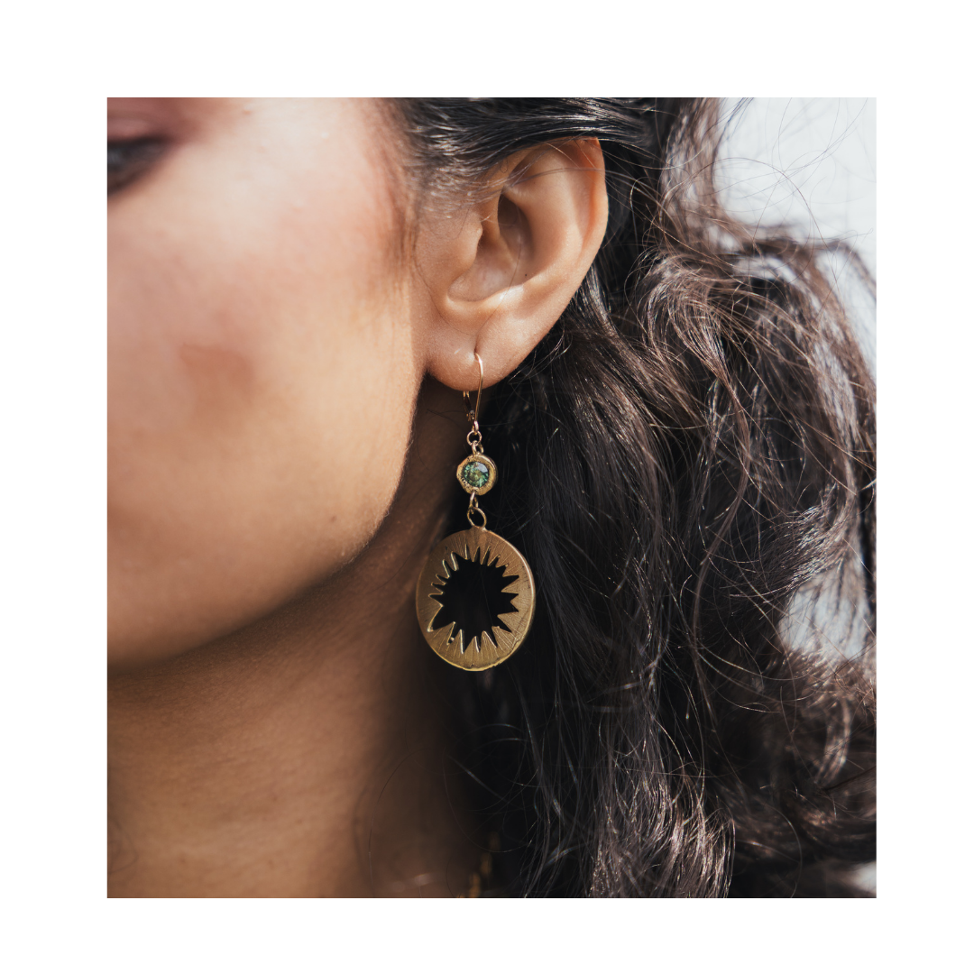 A close-up shows a woman’s wavy dark hair and ear adorned with the Madeleine Blaine Tundra Earrings with Moissanite—round gold earrings featuring a striking black sunburst motif, accented by a small green bead above the pendant. The softly blurred backgro