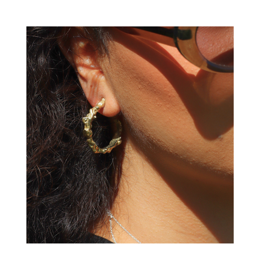 Close-up of a person wearing Molten Hoops by Madeleine Sculptural Jewellery. Their ear, dark curly hair, black sunglasses, and part of a silver sustainable necklace are visible in bright sunlight.