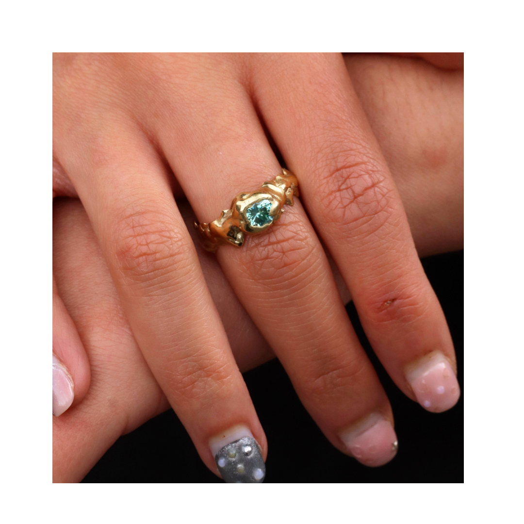 A close-up showcases a hand wearing the Element Ring with Moissanite Small by Madeleine Sculptural Jewellery, featuring green stones. The nails are painted pink, gray, and white, with decorative studs for a handmade look.