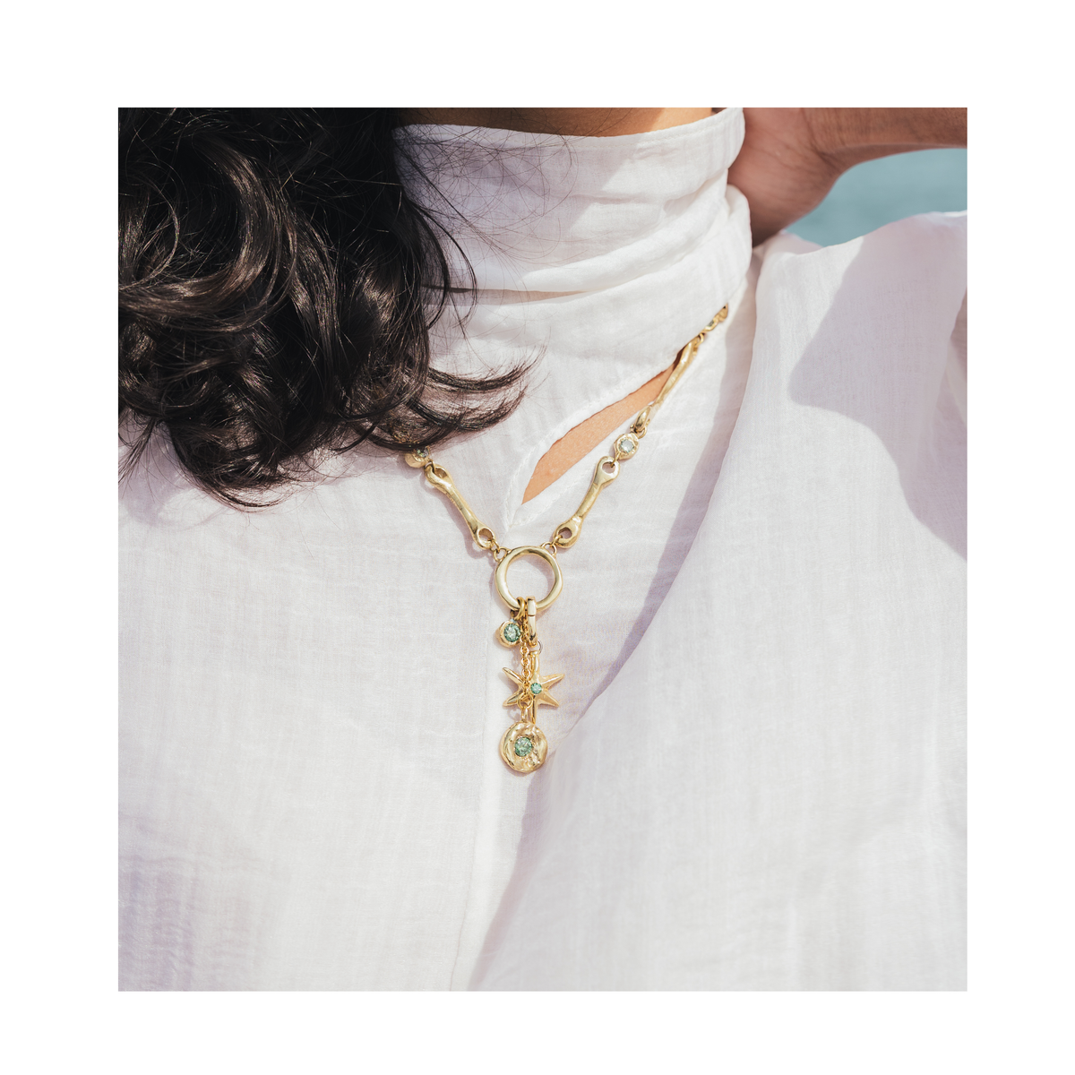 A person wearing a white high-neck top and the Madeleine Blaine Lumen Necklace—a gold handcrafted piece with a circular pendant featuring a star and a green gemstone—shown from the shoulders up.