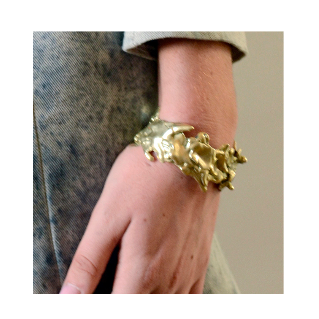 A person wears the Molten Cuff by Madeleine Blaine—a chunky, abstract gold bracelet shaped like a hand—on their wrist. Dressed in a soft gray outfit, their arm rests relaxed at their side, showcasing this striking piece of hand-carved jewelry designed in