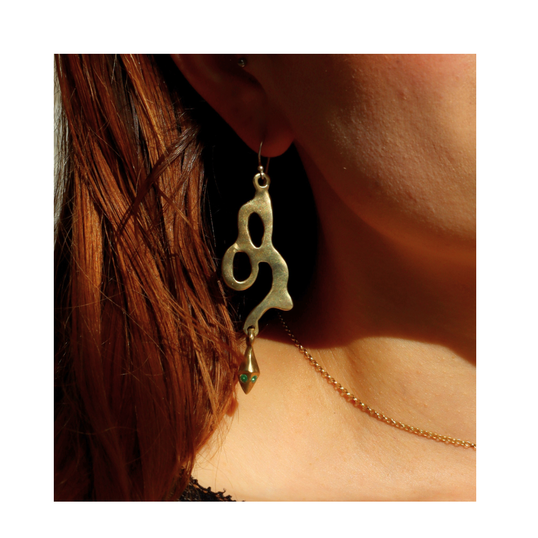 A close-up of a person’s ear adorned with Madeleine Blaine’s Serpent Earrings—large, abstract-shaped gold earrings inspired by serpentine forms. The person, with light brown hair, is also wearing a gold necklace. A plain white background accentuates the u