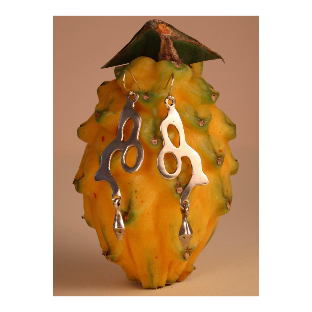 Two abstract Serpent Earrings by Madeleine Blaine, crafted in silver, hang from a yellow dragon fruit with spiky green tips, set against a beige background.