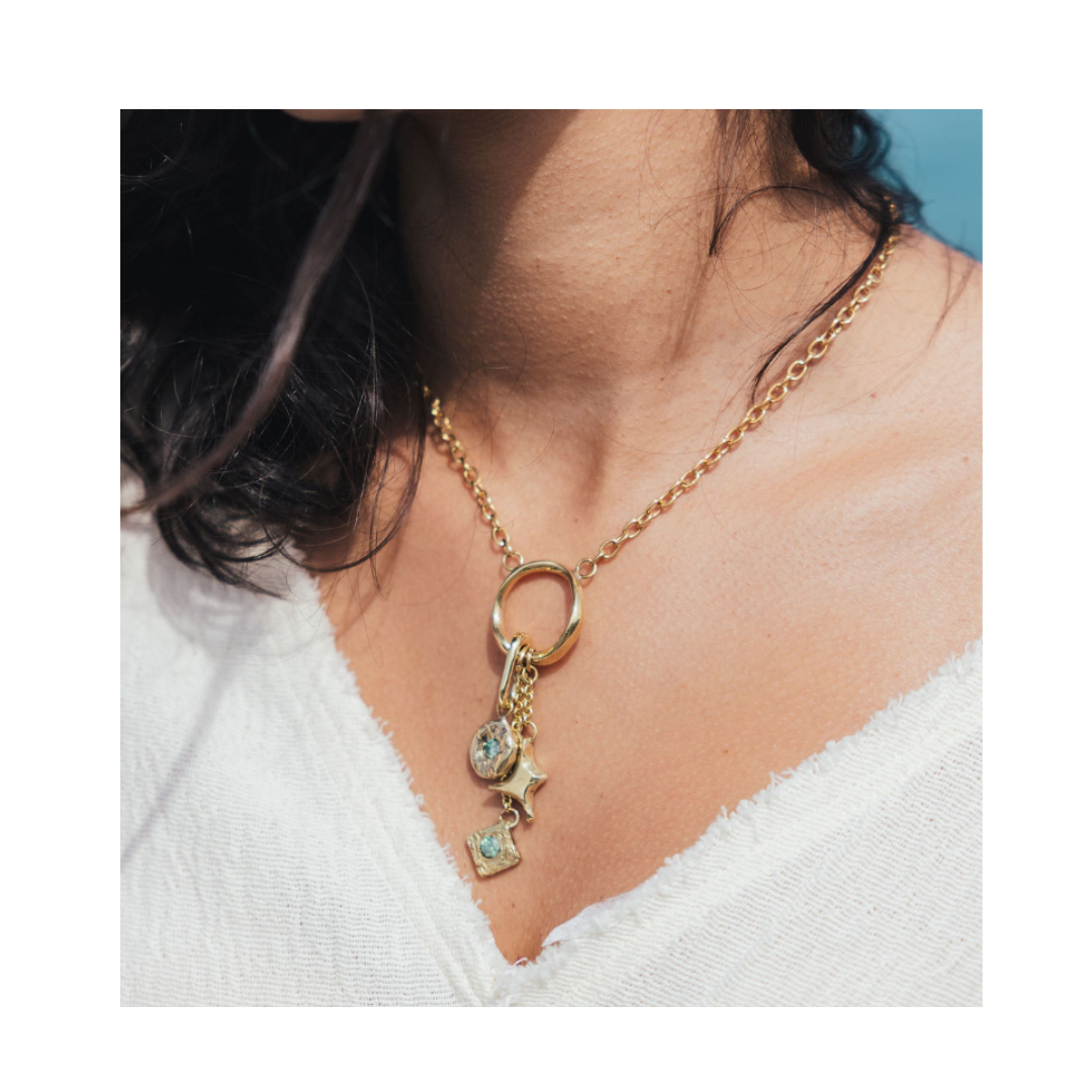 A close-up showcases a woman wearing the Madeleine Blaine Spark Necklace, featuring a gold chain adorned with three delicate charms—a star, a clear gem, and a square gem—over a textured white top. Her dark hair is partially visible as she is captured from