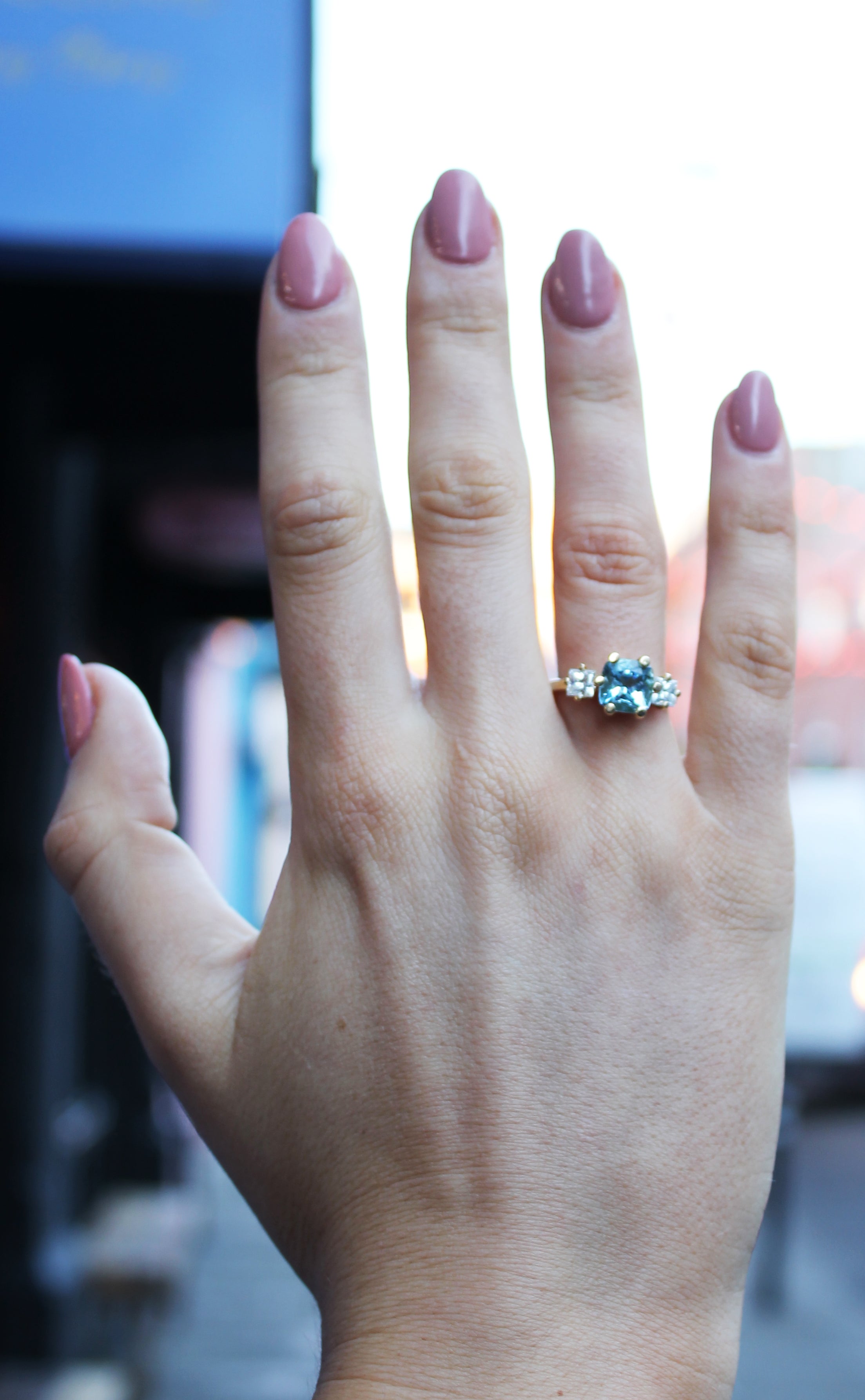 A hand with pink manicured nails displays the Sky's Devotion 18ct Yellow Gold Ring by Madeleine Fine Jewellery—an elegant aquamarine ring with clear accent stones on the ring finger, set against a softly blurred background.