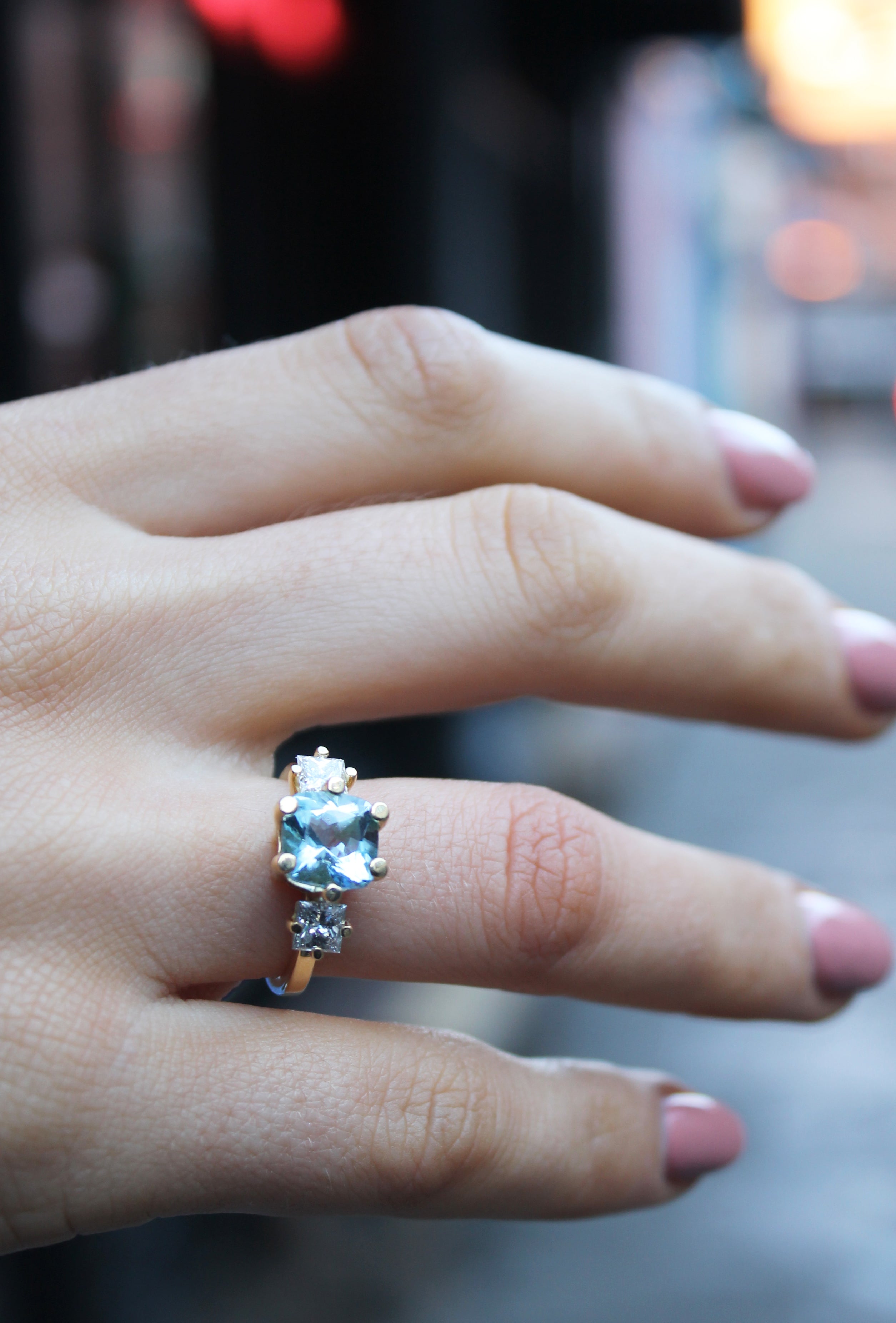 A close-up of a hand with pink nails wearing the Sky's Devotion 18ct Yellow Gold Ring by Madeleine Fine Jewellery, set with three sparkling clear stones, against a blurred outdoor background.
