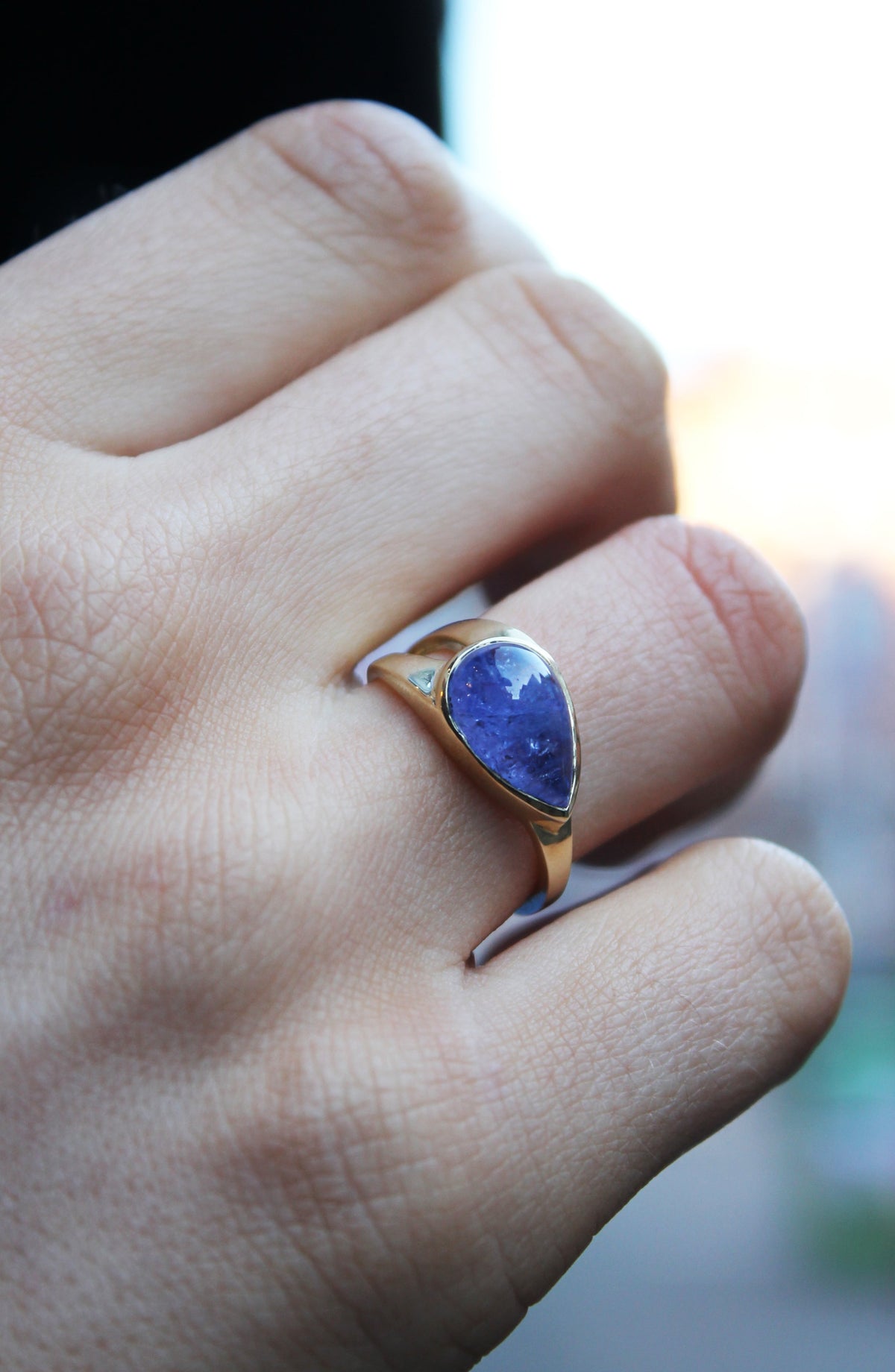 9ct yellow gold ring with teardrop shaped cabochon Tanzanite, which is a dark blue purple stone, bezel set in 9ct yellow gold. Irregular shaped band, tapers from a thicker width with cut-out detail and tapers down to a thinner band. Shown here on a ring f