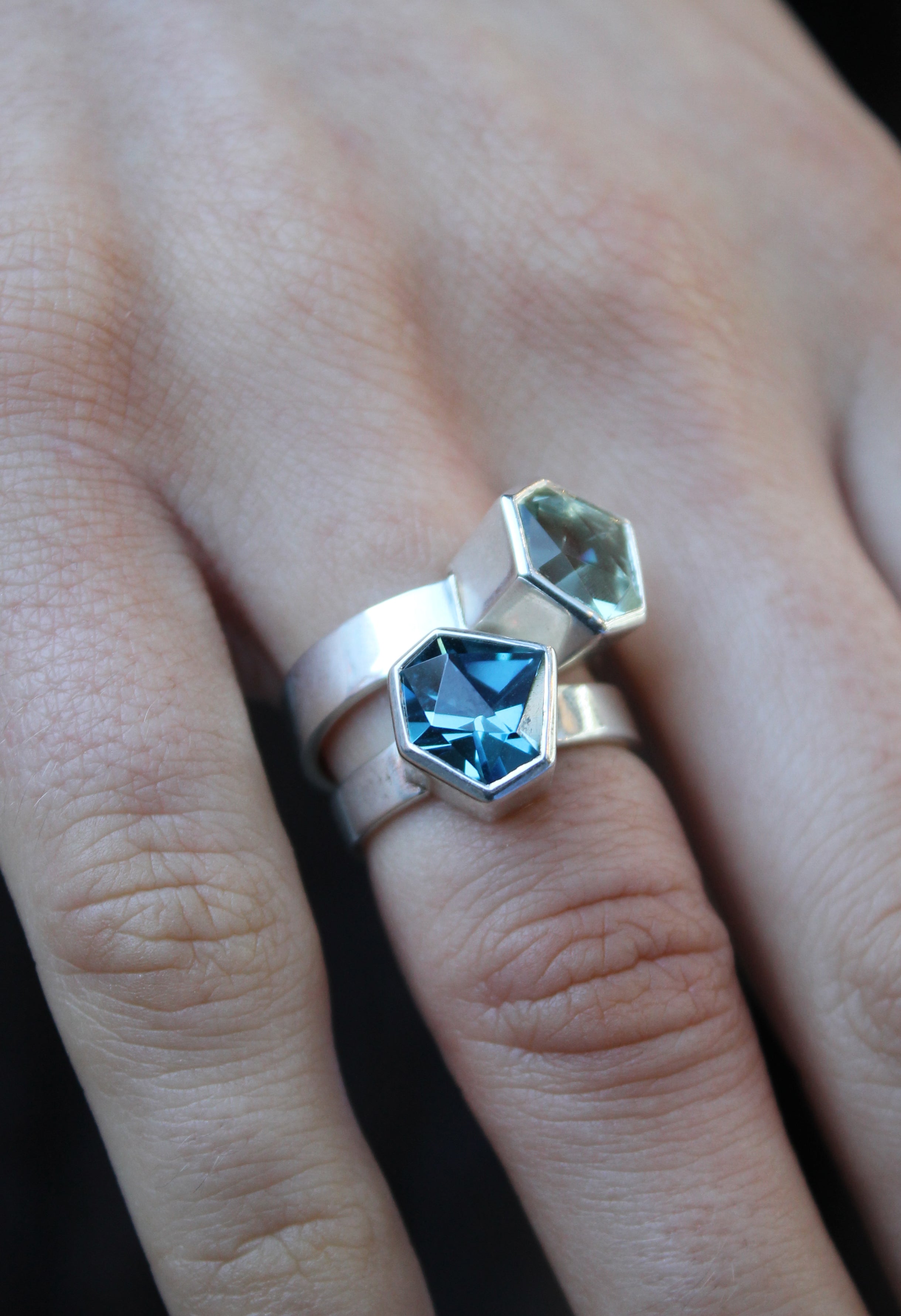 two rings, the first one, closer to the hand is a Silver ring with large, irregular hexagonal green amethyst in a silver bezel. Band is flat and 5mm wide.The second is a Silver ring with large, irregular hexagonal blue topaz in a silver bezel. Band is fla