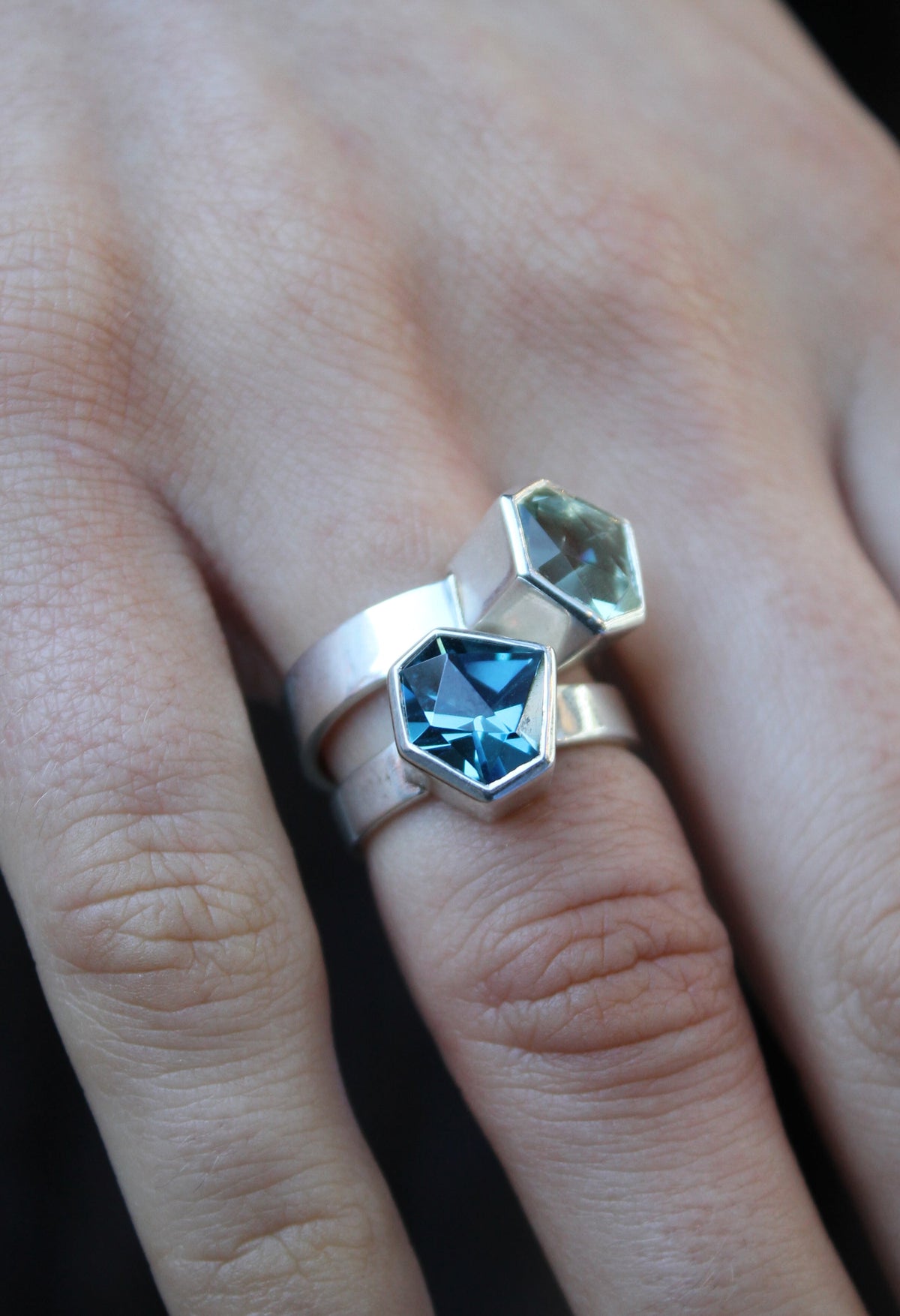 two rings, the first one, closer to the hand is a Silver ring with large, irregular hexagonal green amethyst in a silver bezel. Band is flat and 5mm wide.The second is a Silver ring with large, irregular hexagonal blue topaz in a silver bezel. Band is fla