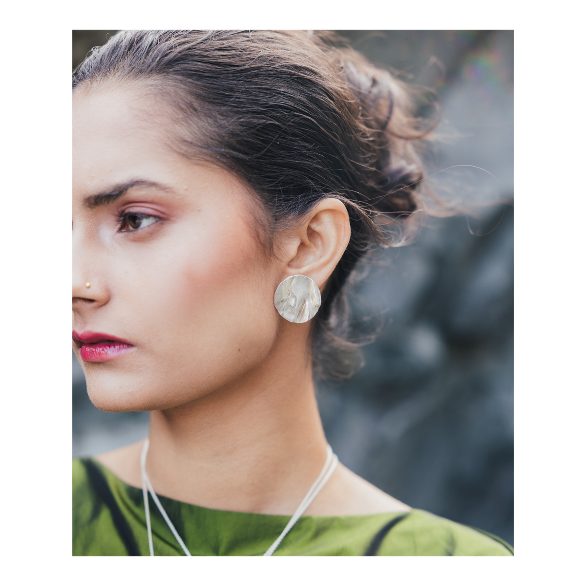 A woman with dark hair styled in a bun wears a large round earring, a nose stud, and a green top. She looks to the left, with a blurred natural background behind her. Her Madeleine Blaine Tide Studs Large—hand-carved jewelry inspired by the rugged beauty