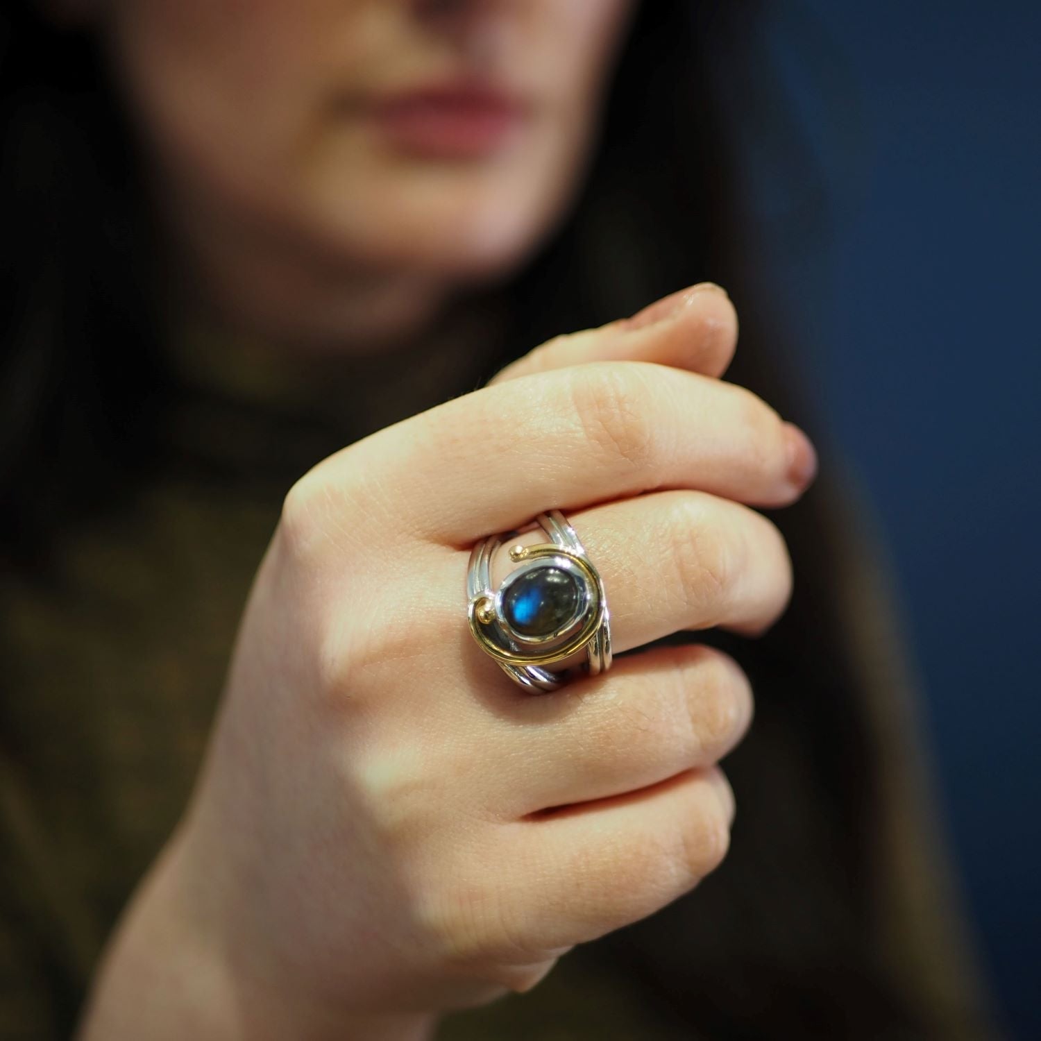 A person with long, dark hair is showcasing a stunning Gallardo & Blaine Designs Jasmine Ring in various gemstones with an adjustable design, featuring a dark, oval gemstone, possibly a blue or black stone. The background is blurred, highlighting the exqu