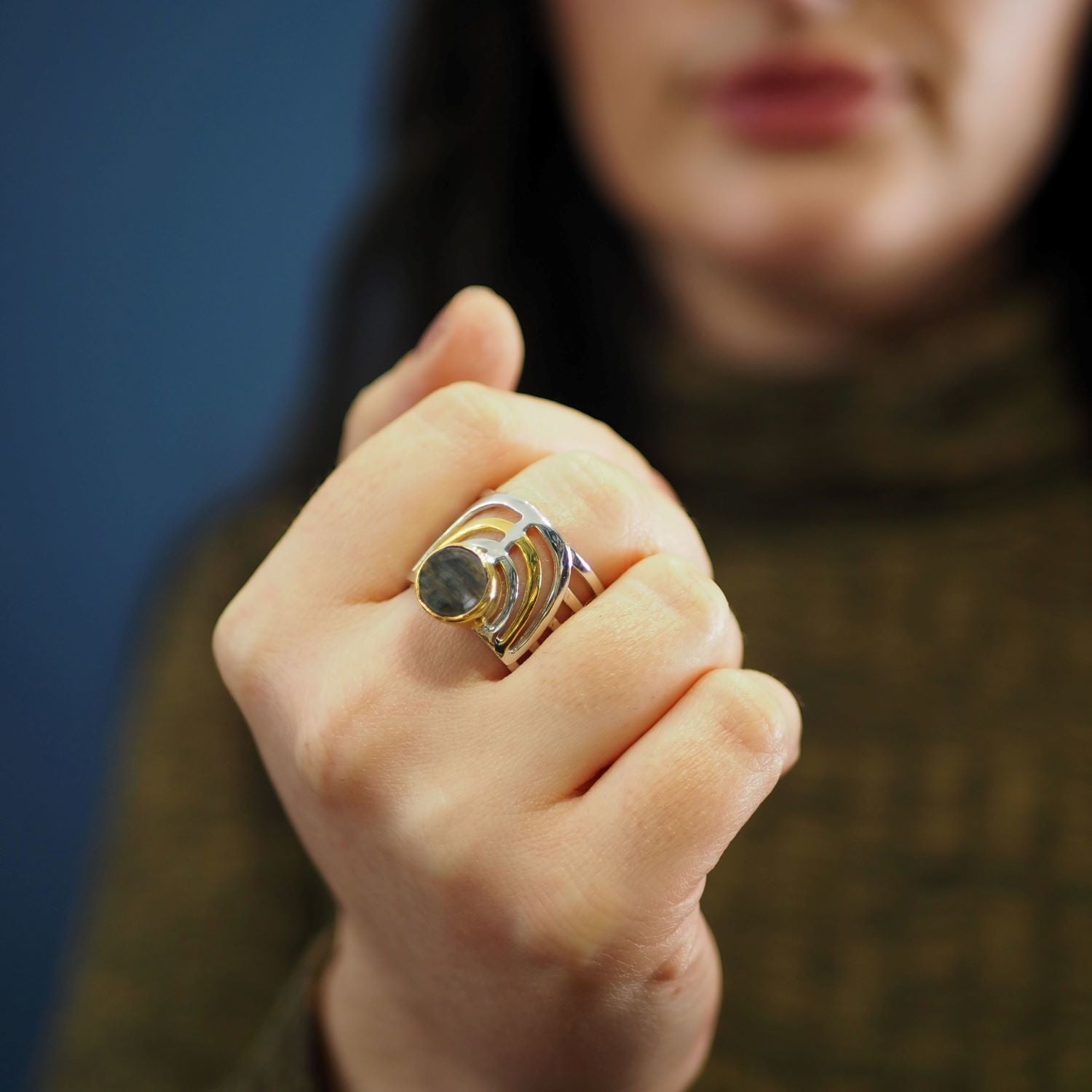 A person with long dark hair wearing a green knit sweater holds up their hand, showcasing a Gallardo & Blaine Designs Solar Ring in various gemstones with a large circular stone and unique design against a blue background. The focus is on the bold jewelle