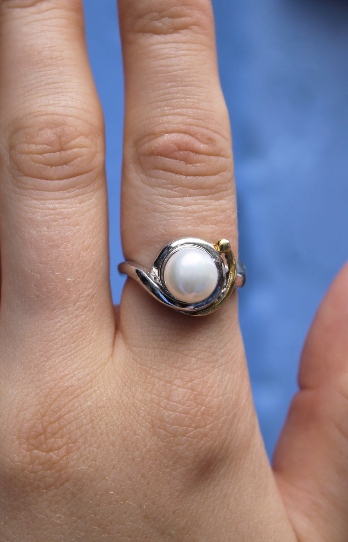 A hand wears Gallardo & Blaine Designs' Wave Ring, featuring a round white pearl in the center with a small gold-plated accent, against a blurred blue background.