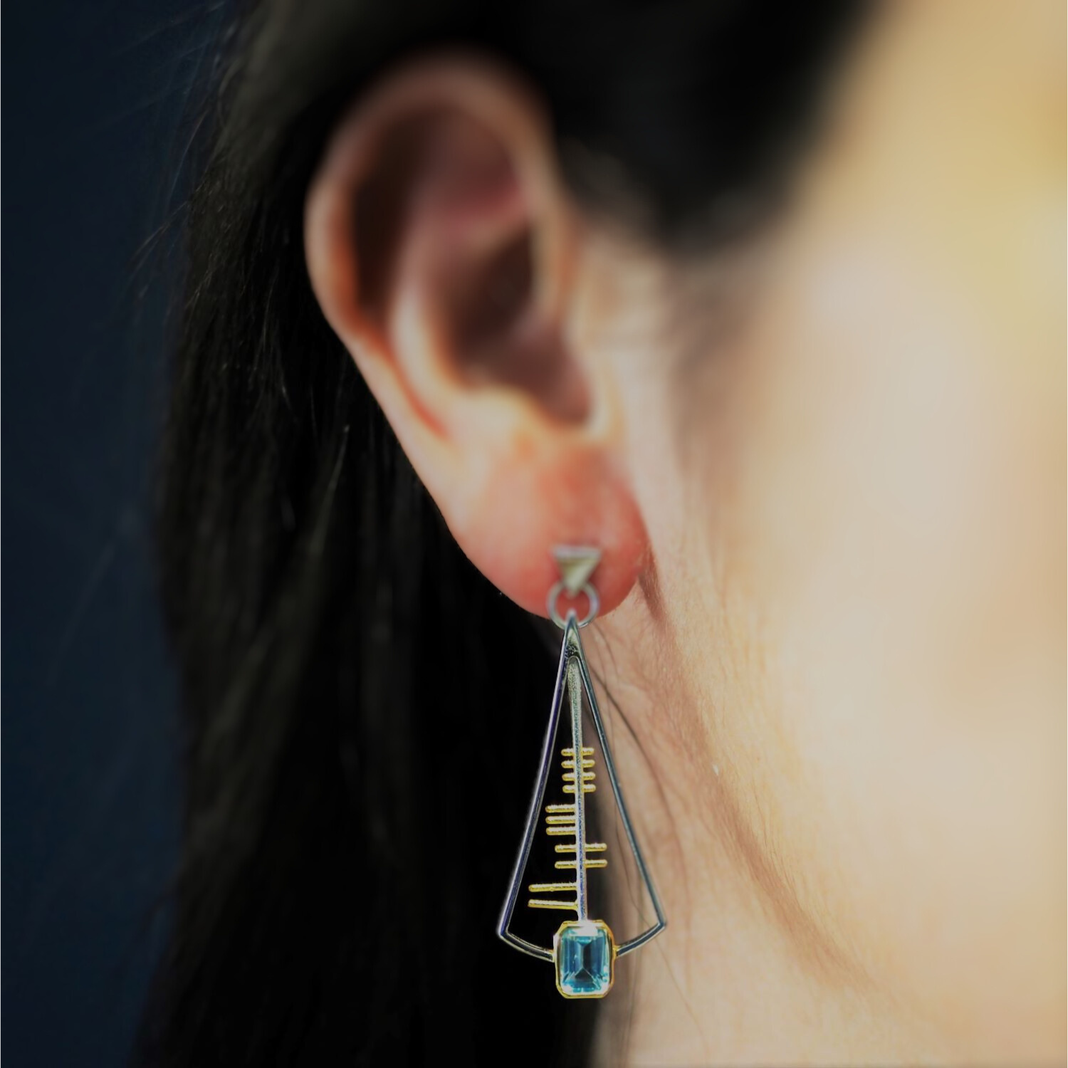 Close-up of a person's ear wearing one of Gallardo & Blaine Designs' Adeline Earrings. The sterling silver triangular frame showcases intricate golden horizontal lines and is adorned with a light blue rectangular gemstone at the bottom. The background is