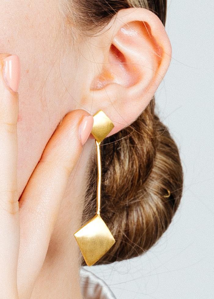 Close-up of a person with a hand touching their face, showcasing an Art Deco earring with a gold vermeil, geometric design from Gallardo & Blaine Designs. The person has light skin and their hair is styled in a low bun. The plain background draws attentio