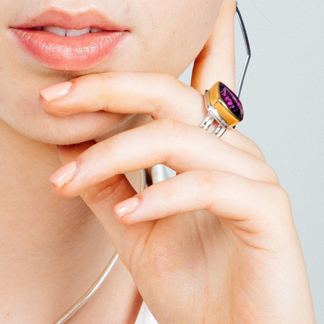 Close-up of a person's face showing their lips and chin, with their hand resting gently on their face. The person is wearing an Art Deco Ring in Silver Gold & various gemstones by Gallardo & Blaine Designs on the index finger and has neatly manicured nail