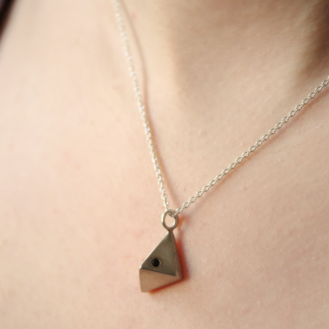 A close-up of the Atria Pendant Necklace by Gallardo & Blaine Designs—a silver chain with a small geometric triangular pendant, adorned with cubic zirconia, resting against bare skin.