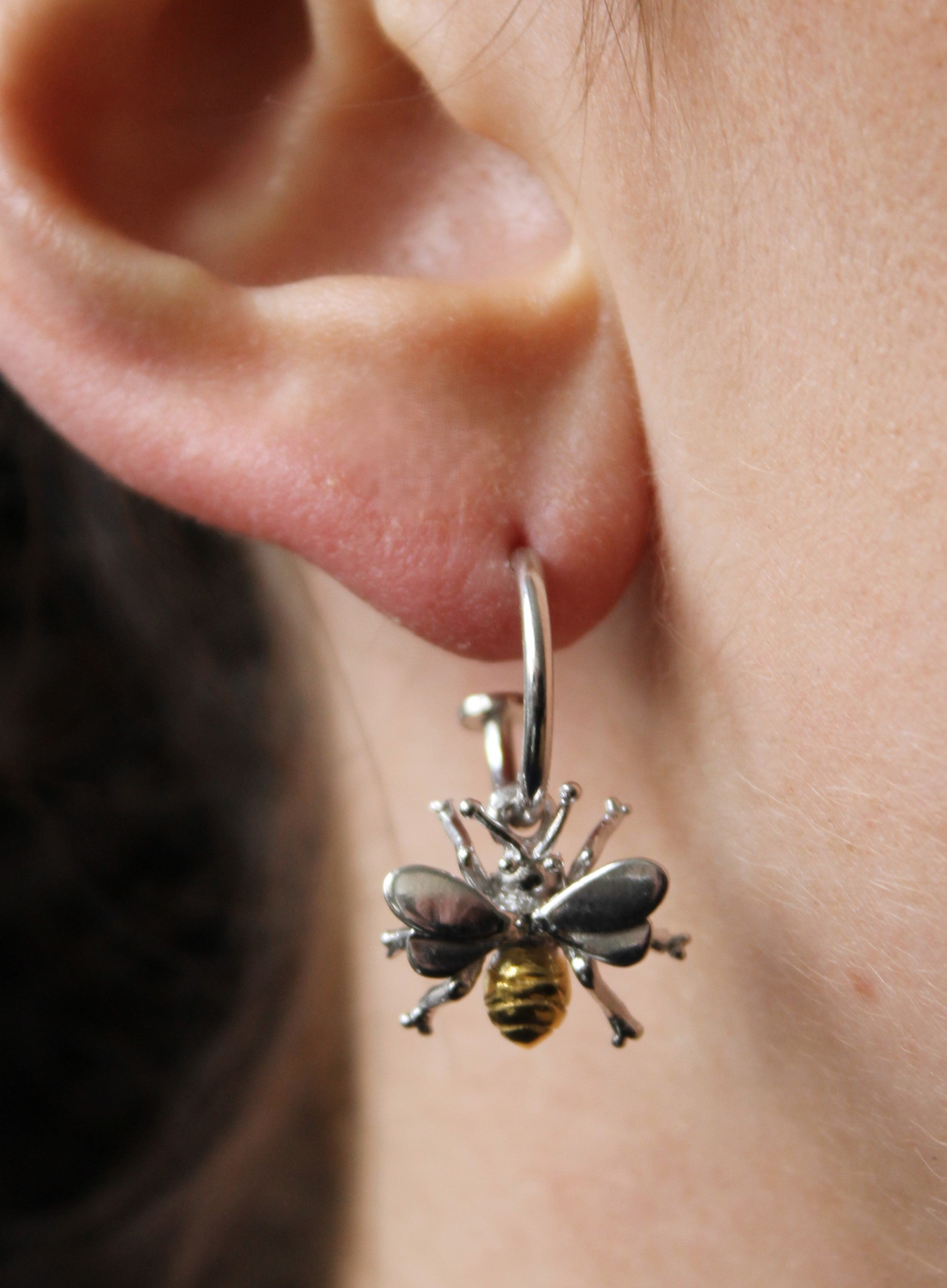 A close-up of a person's ear wearing Watermelon Tropical Wildlife Bee Huggies in silver—a hoop earring with a dangling bee charm featuring silver wings and a yellow striped body, inspired by nature.