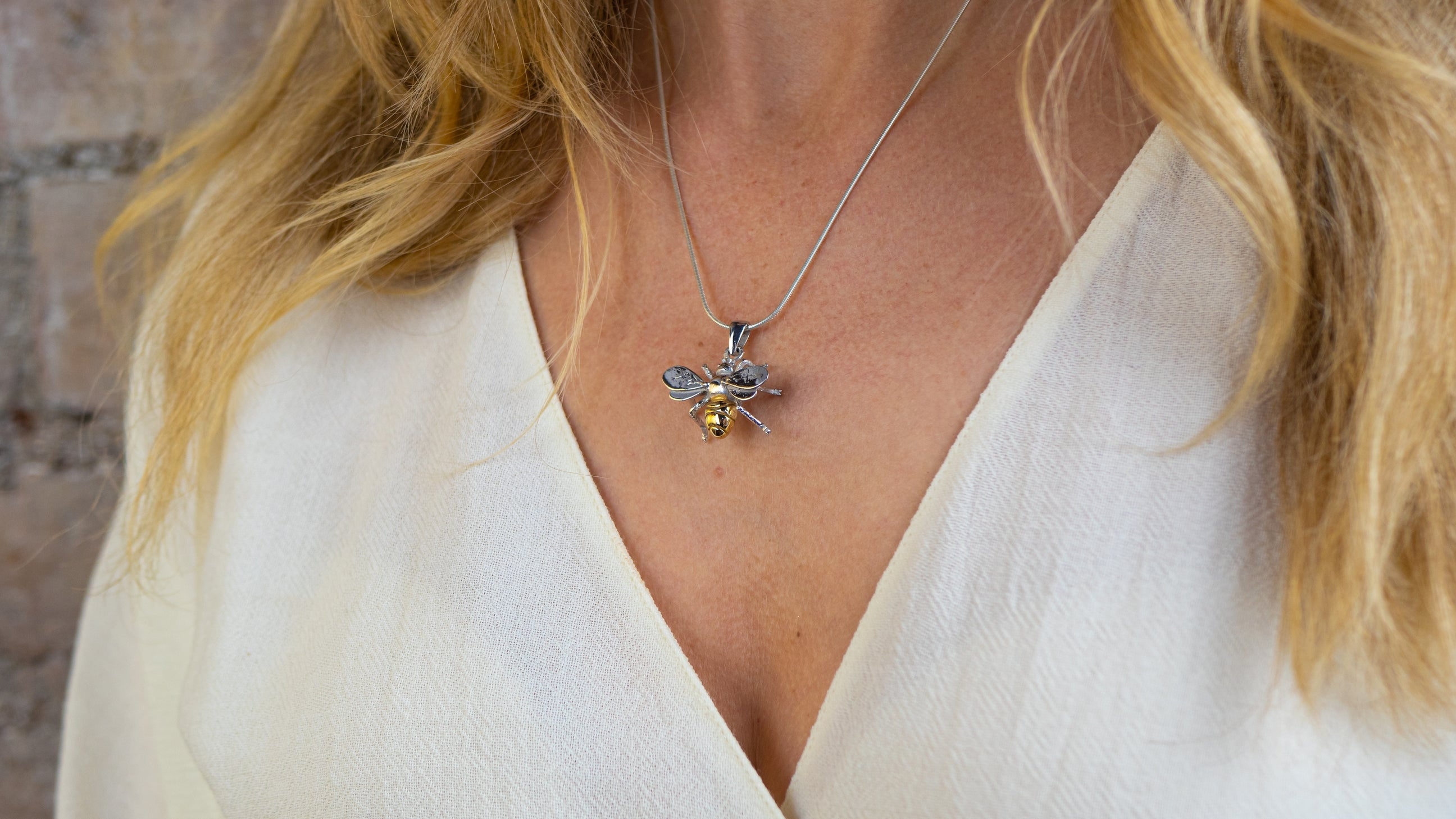 A close-up of a woman in a cream top wearing Watermelon Tropical's Wildlife Pendant - Large Bee With Chain; her blonde hair frames the elegant, nature-inspired gold-plated necklace.