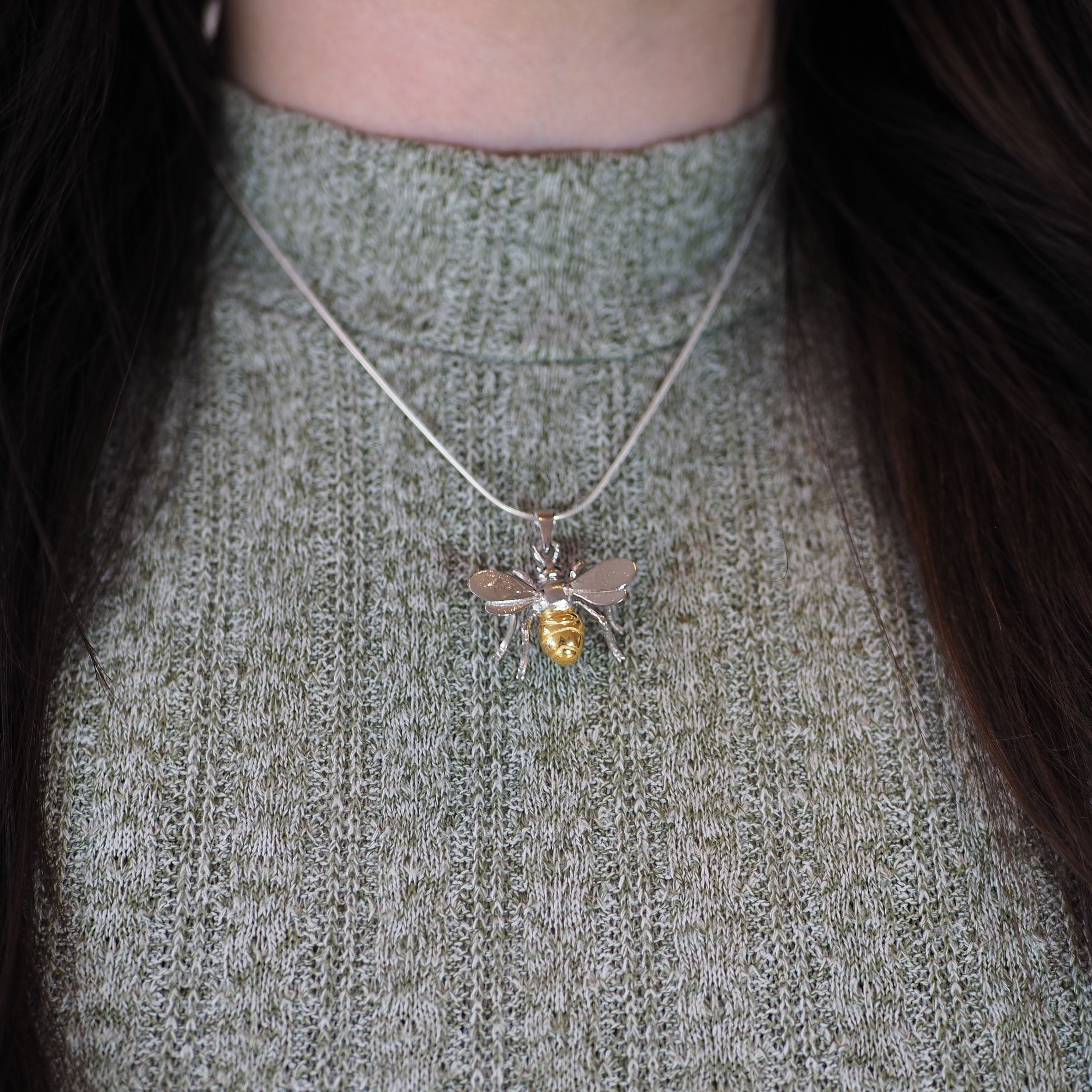 The Wildlife Pendant - Large Bee With Chain by Watermelon Tropical, with its gold-colored body, stands out against a textured green high-neck sweater, while the wearer’s dark hair frames this nature-inspired statement necklace.