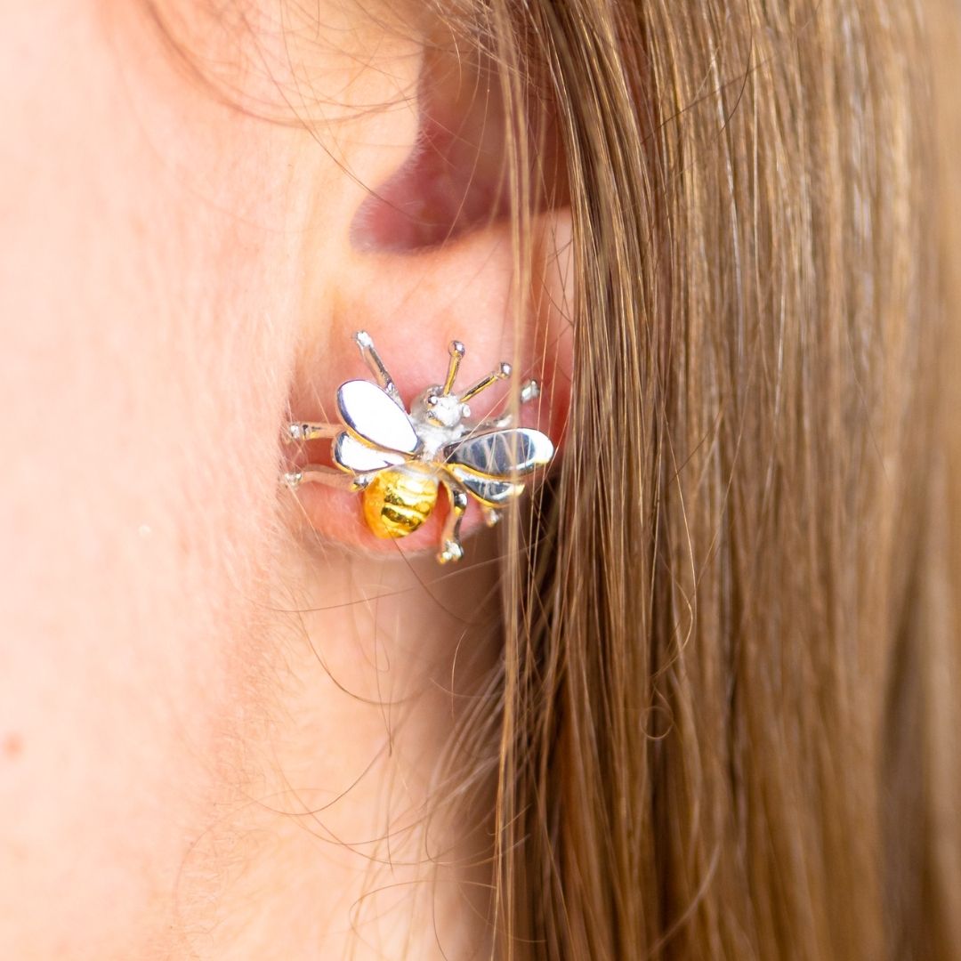 Close-up of a person's ear wearing Watermelon Tropical Wildlife Bee Studs in silver; these nature-inspired earrings with striped bodies and wings add charm, complemented by light brown hair beside the ear.