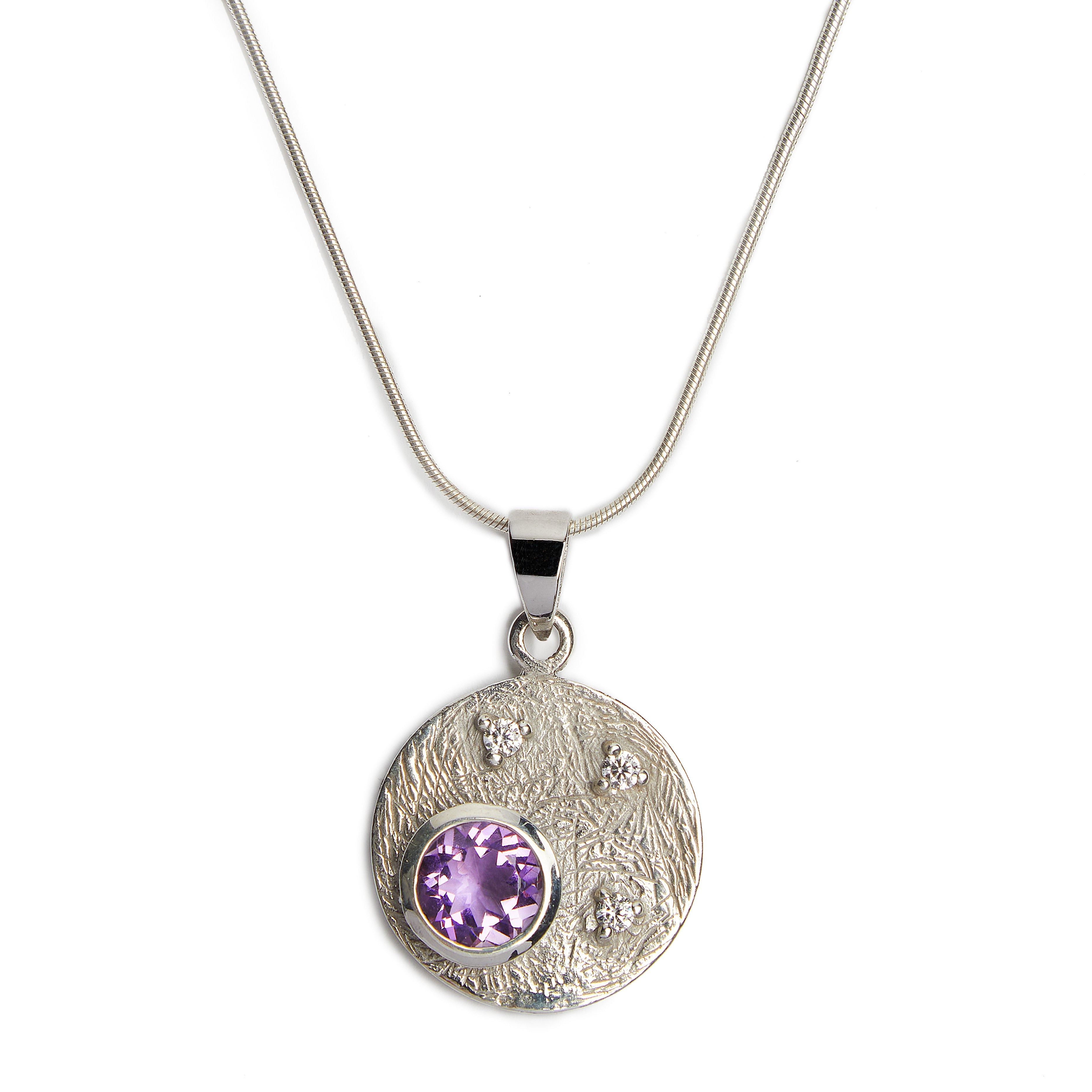 The Gallardo & Blaine Designs Celeste Necklace in Silver & various gemstones is crafted from sterling silver, featuring a round pendant with a textured surface adorned with small cubic zirconia stones and a larger central purple gemstone. The thin, smooth
