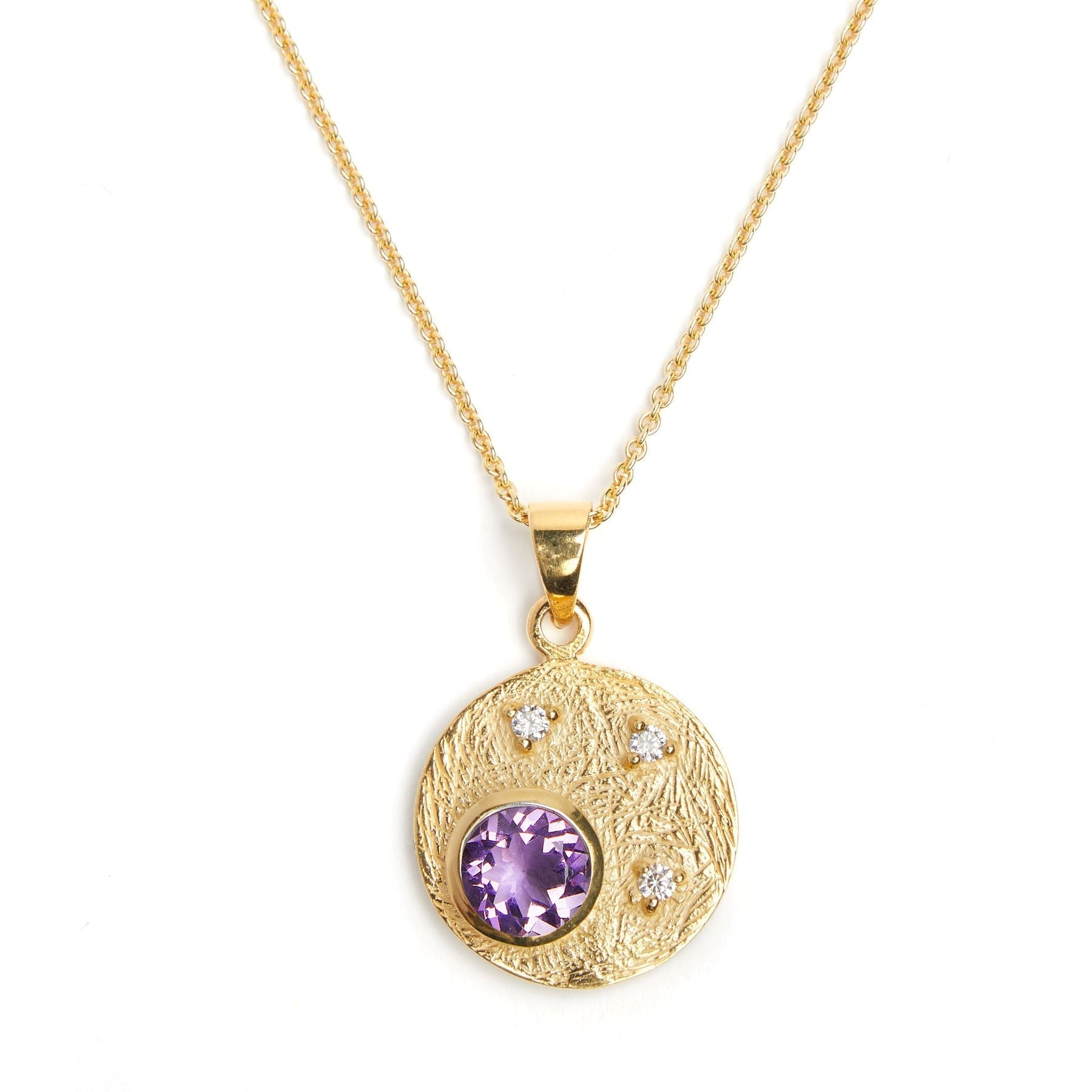 A gold Celeste Necklace in Gold & various gemstones by Gallardo & Blaine Designs featuring a pendant with a textured round disk. The gemstone pendant has a large central purple gemstone and three smaller cubic zirconia stones inset around it. The chain is