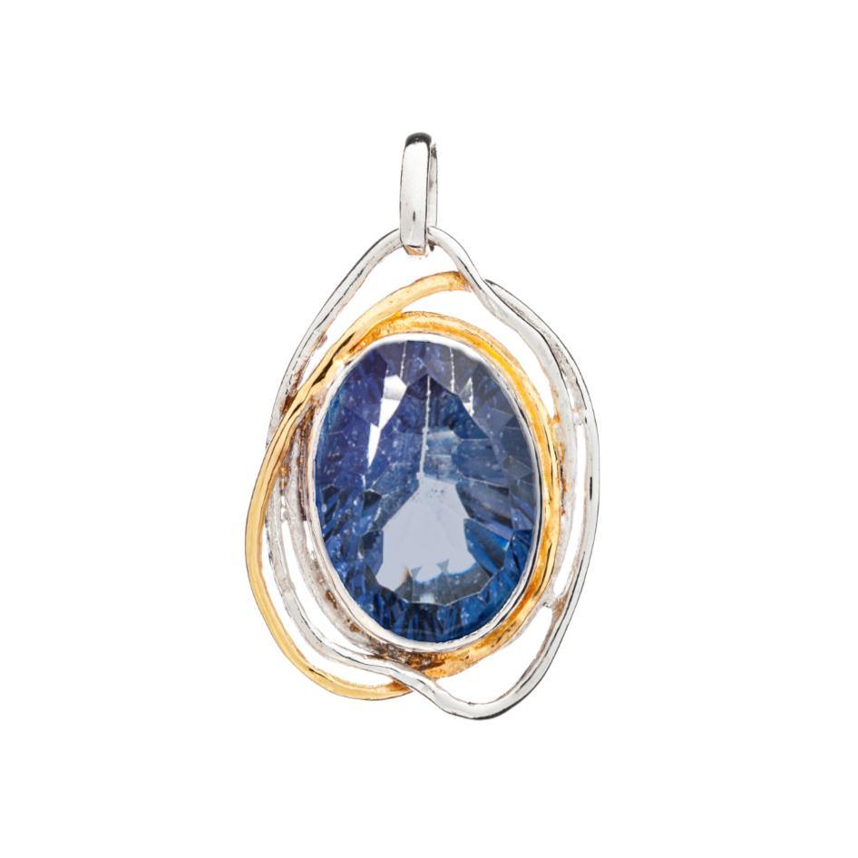 A blue gemstone pendant with a faceted oval sapphire encased in an intricate metal frame featuring silver and gold tones. With its medieval design, the Cosmos Pendant Large in various gemstones by Gallardo & Blaine Designs has a loop at the top for a chai