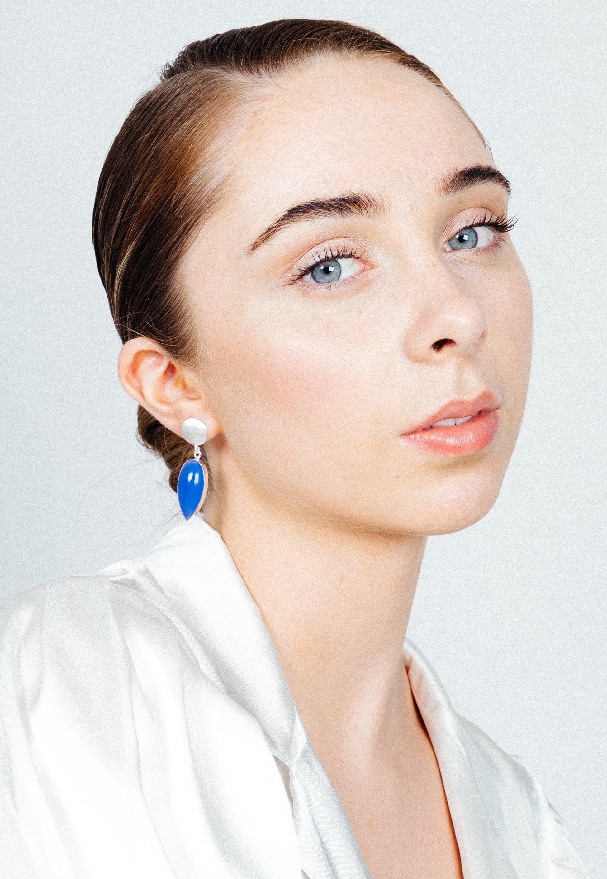 A young person with fair skin, light blue eyes, and brown hair pulled back looks into the camera. They wear a white satin blouse and Gallardo & Blaine Designs' Daira Earrings in sterling silver, featuring a white bead and a blue, elongated teardrop-shaped